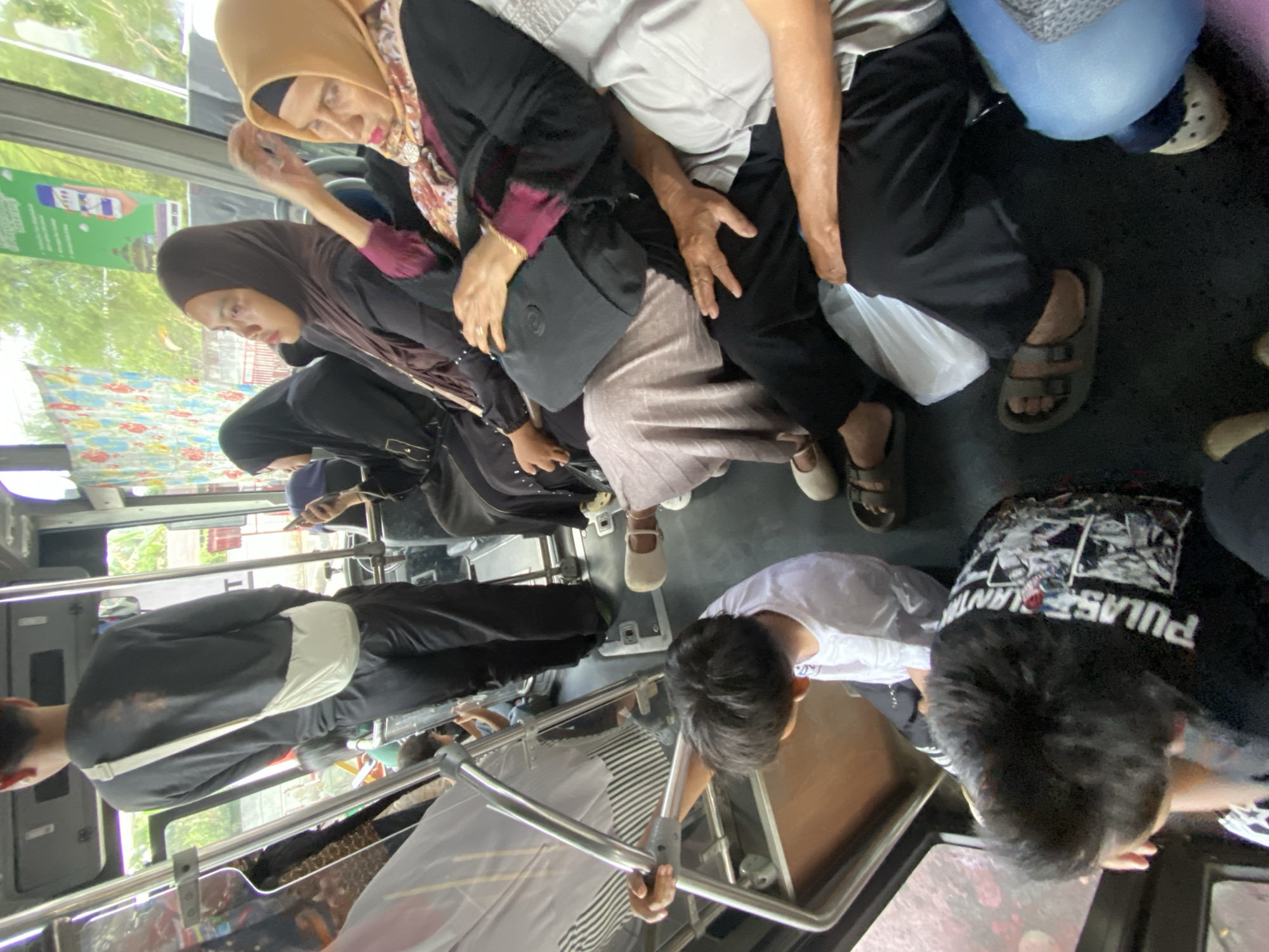 In Banjarbaru, Indonesia, a bus is filled with passengers. A young man stands near the front, wearing a black long-sleeved shirt and black pants, with a white bag slung across his chest. He holds onto a metal pole for support. To his right, a group of women are seated. The woman in the foreground wears a yellow hijab and a black outfit with a maroon detail. Next to her, another woman is dressed in a dark hijab and black clothing. Further down the aisle, more passengers are visible. A young boy in a white shirt sits with his back to the camera, holding onto a metal railing. Another passenger in a black shirt with a graphic print is seated in the foreground. The interior of the bus is visible, with windows showing a blurred outdoor scene of trees and buildings. The floor is dark, and several people are wearing sandals.