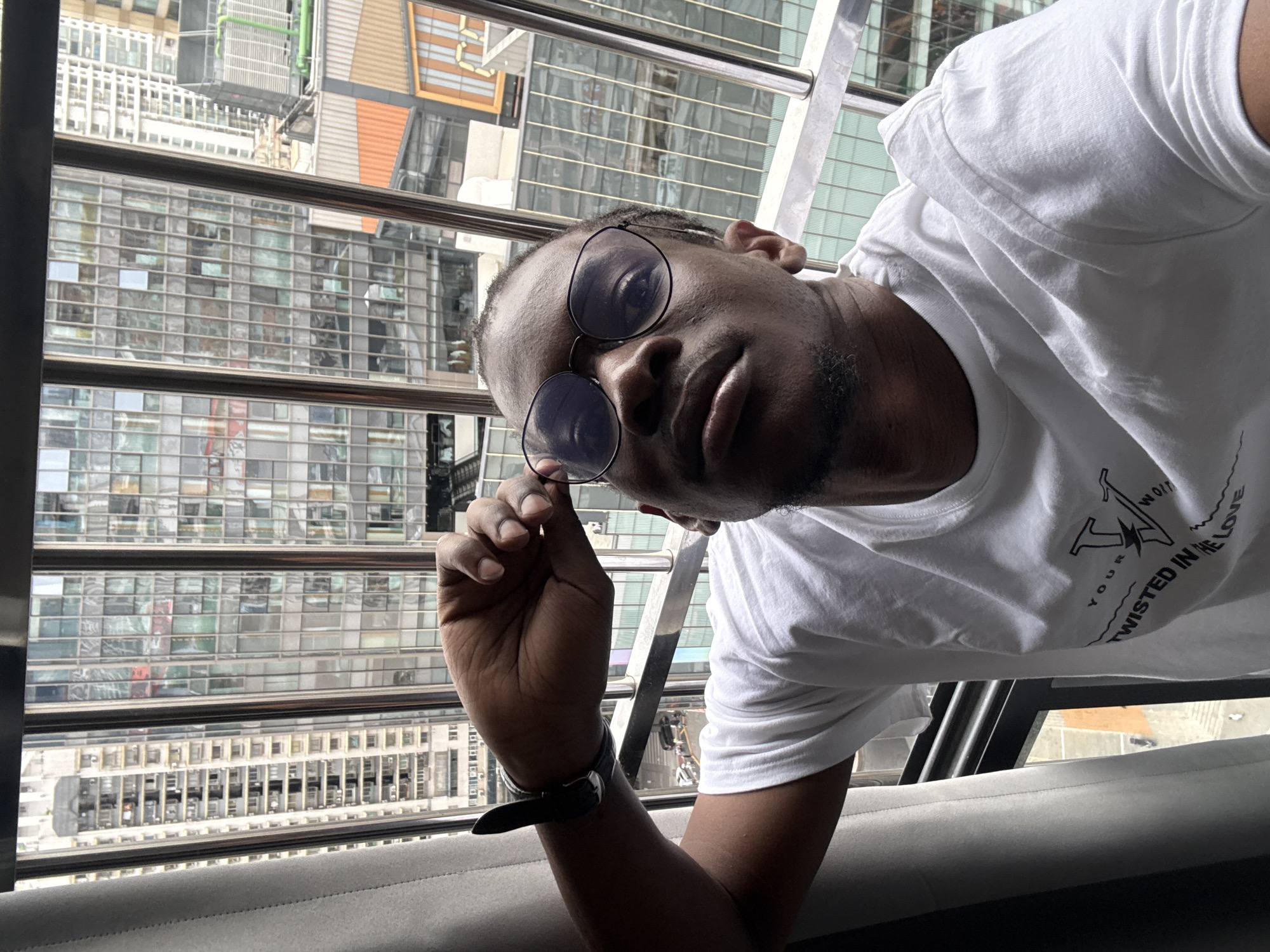A young Black man wearing a white t-shirt and round glasses is looking upwards and slightly to the left, with his right hand holding the side of his glasses. He is positioned in front of a large window that offers a view of a dense urban cityscape with many tall buildings. The man has short dark hair and a goatee. His t-shirt has a graphic print on the chest area. He is wearing a dark watch on his left wrist. The overall environment suggests an indoor setting with a view of a city, possibly Yuzhong District, China, given the architecture.