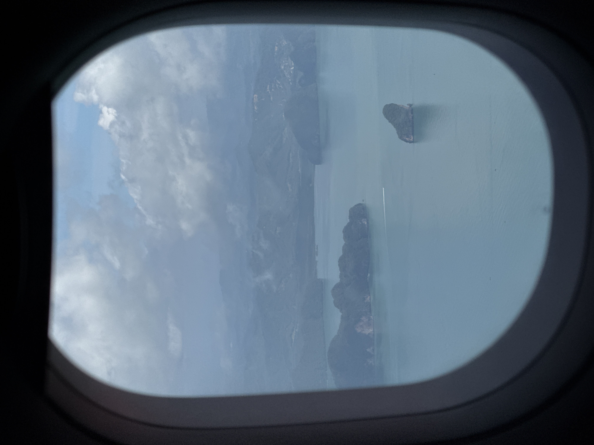 An aerial view is captured through an airplane window, looking out over the coastal landscape of Laem Sak, Thailand. The scene is dominated by the calm, turquoise waters of the sea, dotted with several lush, green islands. The islands vary in size, with some appearing as small rocky outcrops and others as larger landmasses with trees covering their slopes. In the distance, hazy mountains rise, partially obscured by a layer of clouds. The sky above is a mix of blue and white, with fluffy clouds creating a dynamic pattern. A thin, white streak of a boat is visible on the water, suggesting movement and activity in the otherwise serene environment. The overall impression is one of natural beauty and tranquility, characteristic of the tropical scenery of the region.