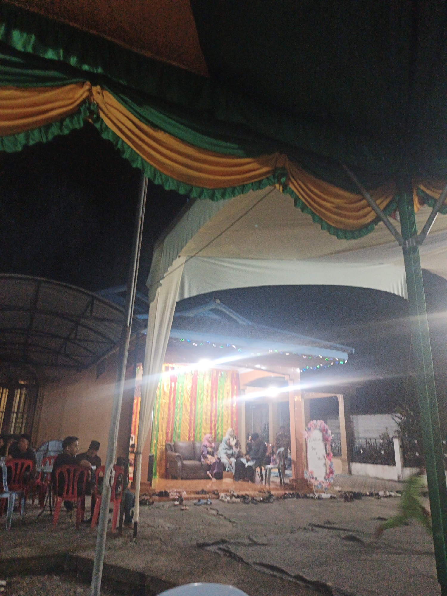 The image shows an outdoor event, likely a reception or gathering, taking place at night under a tent. The tent is decorated with yellow and green fabric draped along the top edge. In the background, there is a stage area with colorful vertical drapes, a brown sofa, and people seated. There are also several rows of red and blue chairs set up in the foreground, with people sitting in some of them. A large sign with floral decorations stands to the right of the stage. The ground appears to be covered in dirt or sand. The presence of a tent and the arrangement of seating suggest a community event. The location is identified as Pematang Duku, Indonesia, indicating a local cultural or social gathering.