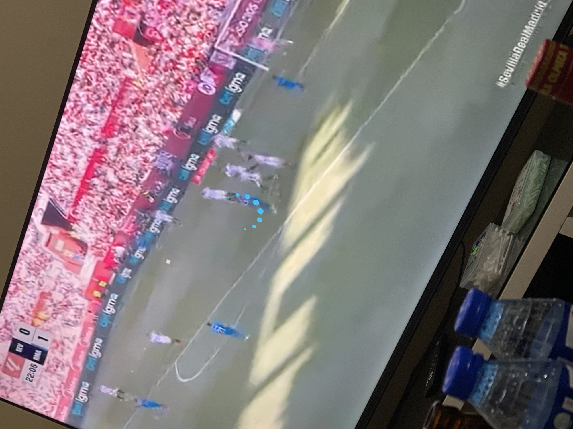 The image shows a television screen displaying a soccer game. The score is Sevilla 0, Real Madrid 1, and the time is 22:06. The game is being played in front of a large crowd. Text such as "belgma" and "Socios" are visible as advertising. The screen also displays "#SevillaRealMadrid." In the foreground are two water bottles with blue caps and a small bottle of a brown liquid. There is also a container of cotton swabs, and a red bottle with a label partly showing. The context lacks any information about the city of Abuja, Nigeria.