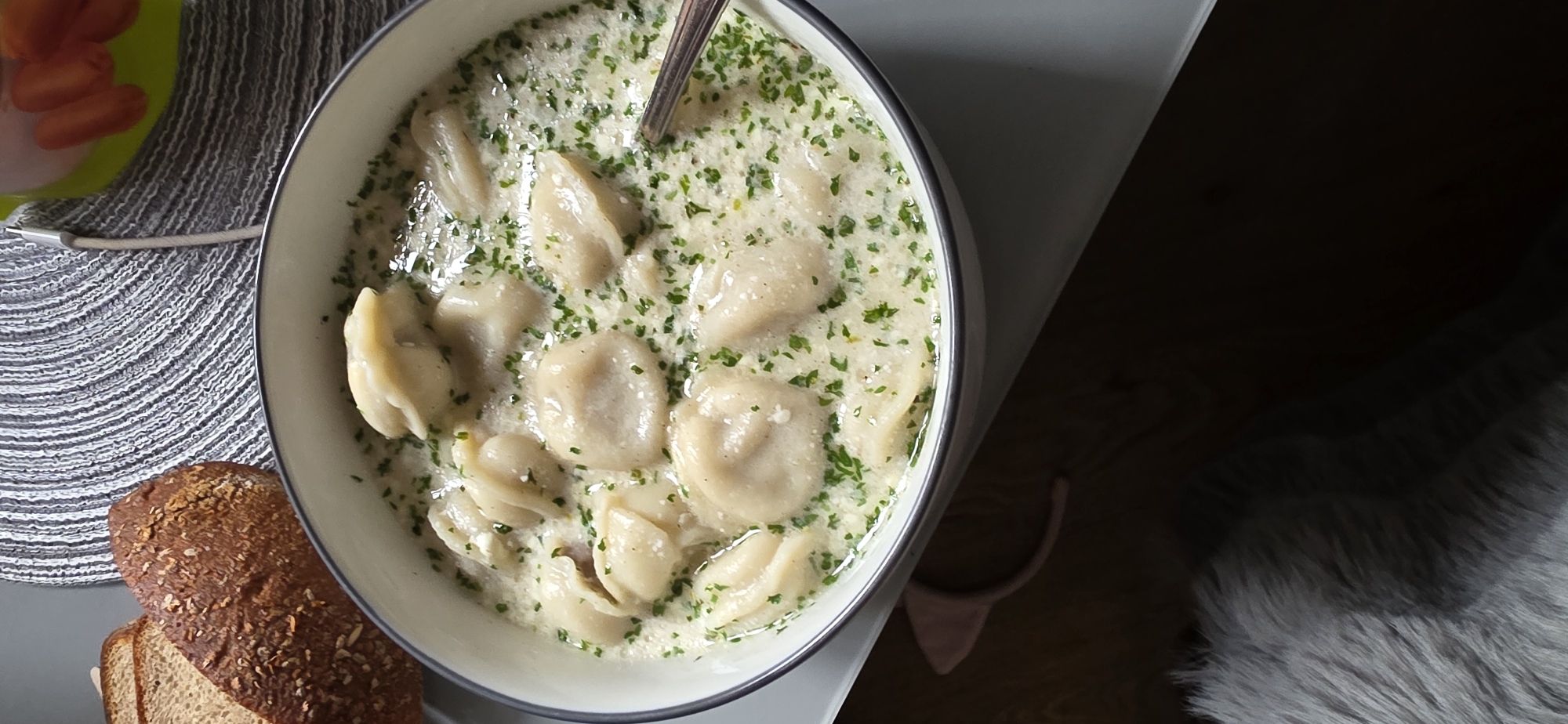 A close-up, overhead view shows a white bowl filled with dumplings in a creamy sauce and chopped parsley. To the left of the bowl are two slices of dark, textured bread. The bowl is placed on a grey placemat with a patterned design. In the background, a portion of a green container with what appear to be orange food items is visible, along with a charging cable. The overall setting suggests a meal being served on a table. There are no identifiable landmarks or features that would place this scene in Arun, United Kingdom.