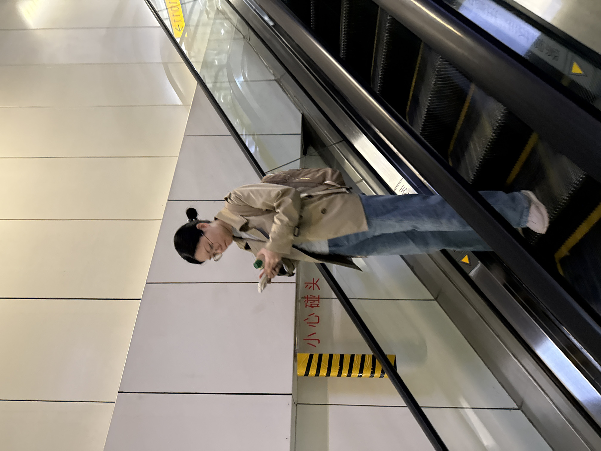 A young woman wearing glasses and a tan trench coat, blue jeans, and white shoes stands on a moving escalator in an urban environment. She is looking down at a small green object in her hands. A sign in red Chinese characters is visible next to her, and a yellow and black striped safety bumper is below it. The escalator is framed by stainless steel. The location is likely in the Yuzhong District of China.