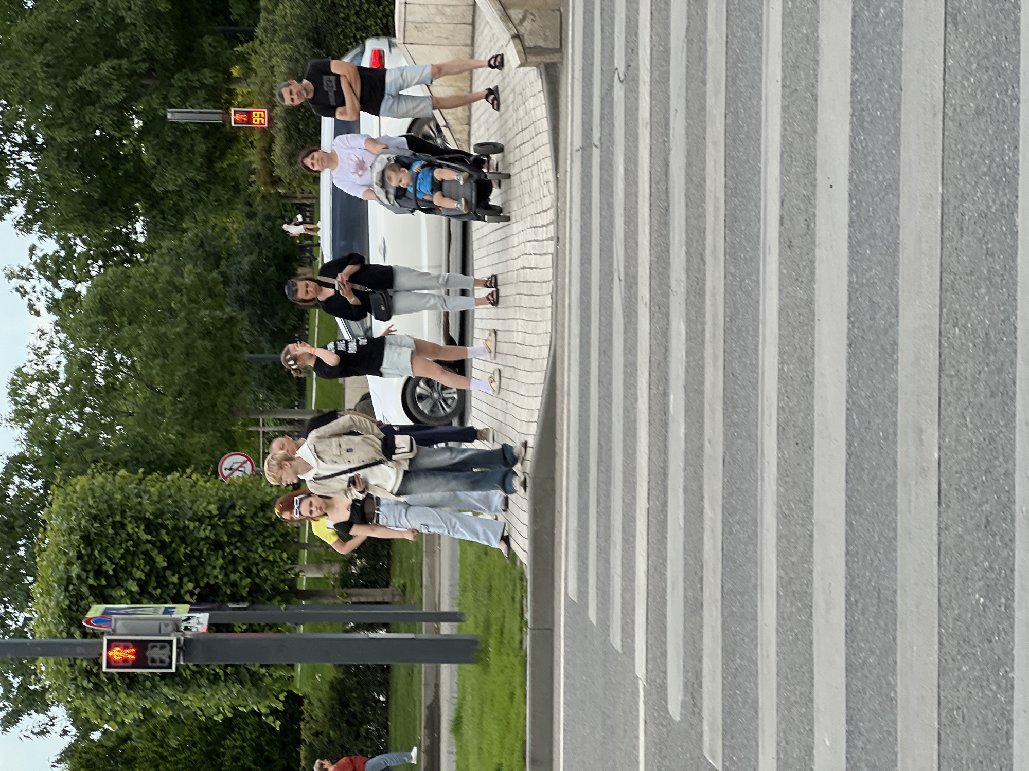 A group of people is captured waiting at a pedestrian crossing during the daytime in Krasnodar, Russia. The image is oriented sideways, but when viewed upright, a wide zebra crossing with alternating grey and white stripes is prominent on the right side of the frame. On the left, a diverse group of approximately seven adults and one child in a stroller are gathered on a paved sidewalk, poised to cross the street.
Among the group, a person in a white top is pushing a black stroller containing a small child dressed in blue. Further along the line, other adults are standing, some engaged in conversation or looking towards the street. To the far left, a traffic light pole stands tall, displaying a red pedestrian signal icon and the number "66," indicating a countdown for not crossing. The background is filled with lush green trees and foliage, suggesting a park-like or tree-lined urban environment. A white car is partially visible behind some of the individuals. The scene portrays a common urban moment of people patiently waiting for the traffic signal to allow them to proceed across the road. No strong emotions are discernible, with individuals appearing calm and observant.