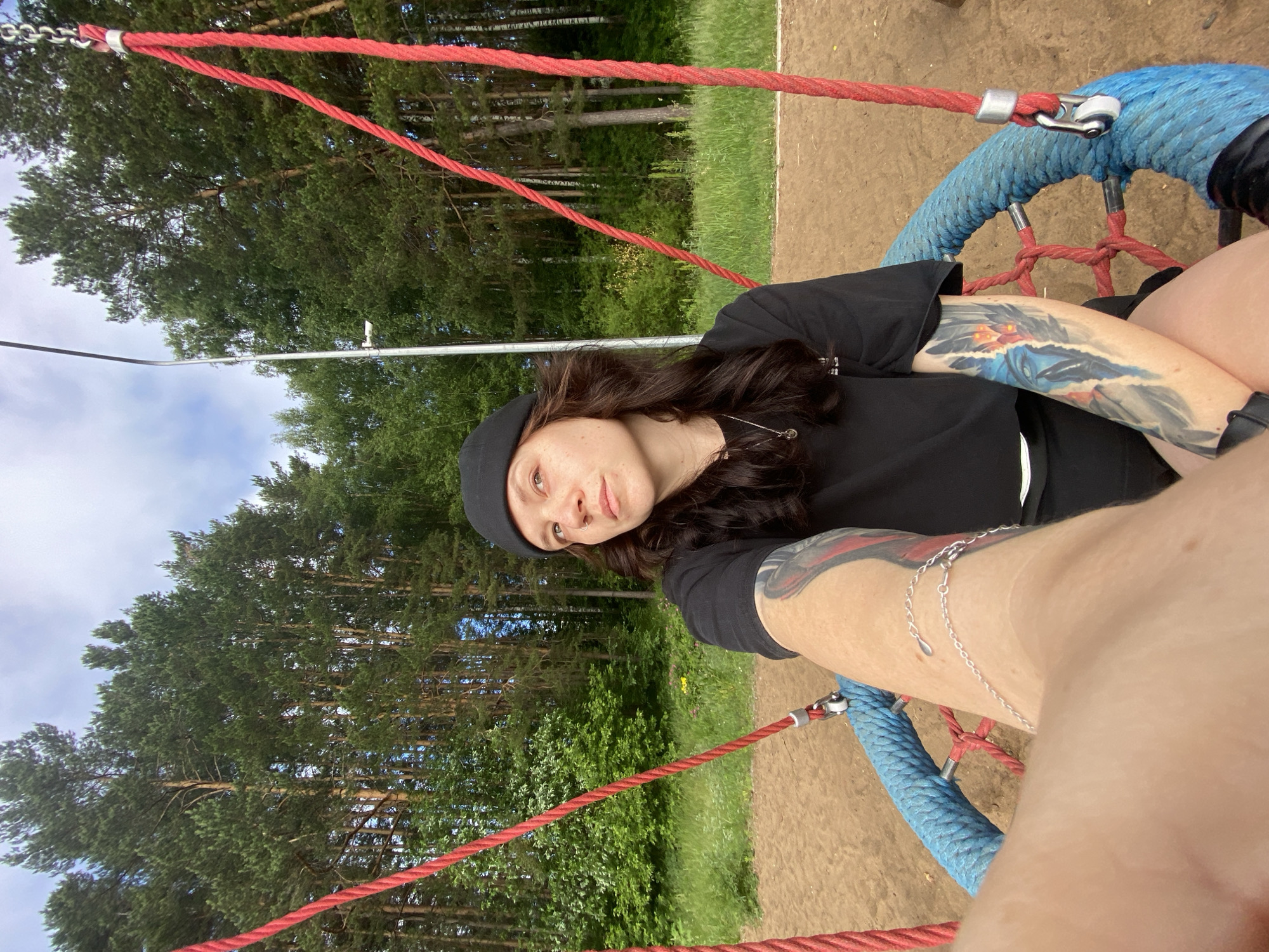 A young woman with dark, wavy hair, wearing a black beanie and a black t-shirt, sits on a round blue swing with red ropes, suspended outdoors. She has tattoos on both arms, the visible one on her right arm depicting a blue and orange feathered design. The swing is set in a sandy area, with a background of dense green pine trees under a cloudy sky. A metal pole with a light fixture is visible to the left of the swing. The overall scene suggests a park or recreational area within the city of Chaikovsky, Russia, given the style of the trees and general environment.