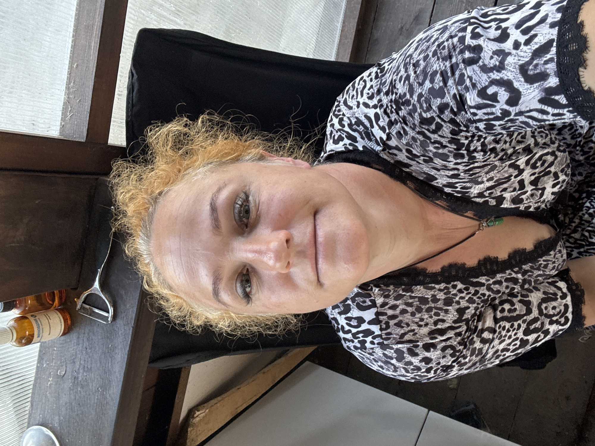 A medium shot shows a blonde woman with curly hair, wearing a leopard print top with black lace trim, and a dark necklace with a green pendant. She is seated in a chair in what appears to be a greenhouse or sunroom. In the background, there is a wooden shelf with two small bottles and a peeler. The overall lighting suggests it is daytime. There is no indication this image was taken in Chaikovsky, Russia.