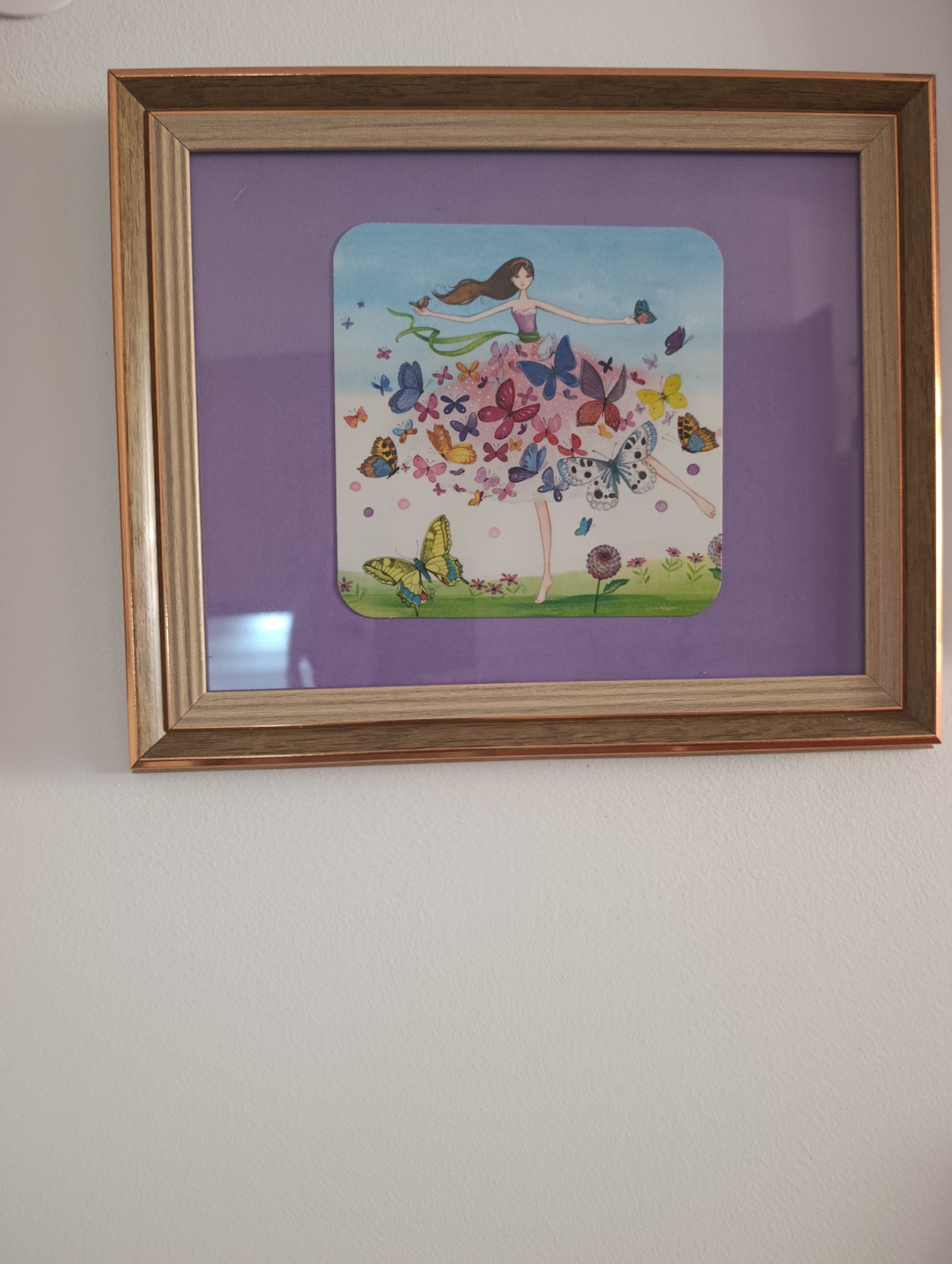 A framed watercolor painting of a young girl with long brown hair wearing a pink dress is displayed on a white wall. The girl is depicted in a whimsical pose, surrounded by a multitude of colorful butterflies. The painting is matted with a lavender-colored mat and framed with a wooden frame that has a metallic copper finish. The painting is hung at eye level. The surrounding environment is a plain white wall. There is no discernible information in the image to indicate its location in Setúbal, Portugal.
