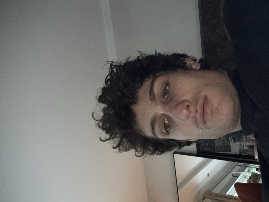 A male individual is depicted in a close-up, slightly upward-angled view within an indoor setting. He has dark, curly hair and light-colored eyes, which are directed downward and to the viewer's left. His nose has a slight reddish tint at the tip, and he exhibits subtle stubble on his face. He is wearing a dark, high-necked garment, likely a turtleneck. The upper portion of the image shows a plain, light-colored ceiling. To the left of the subject, a large, dark-framed mirror reflects a window with vertical elements, and a hint of an orange-red soft furnishing is visible. The right side of the background shows a dark, textured surface, possibly part of a furniture piece. The lighting is even, suggesting ambient indoor illumination. The specific location within Vincennes, France, cannot be determined from the visual information presented in the image.