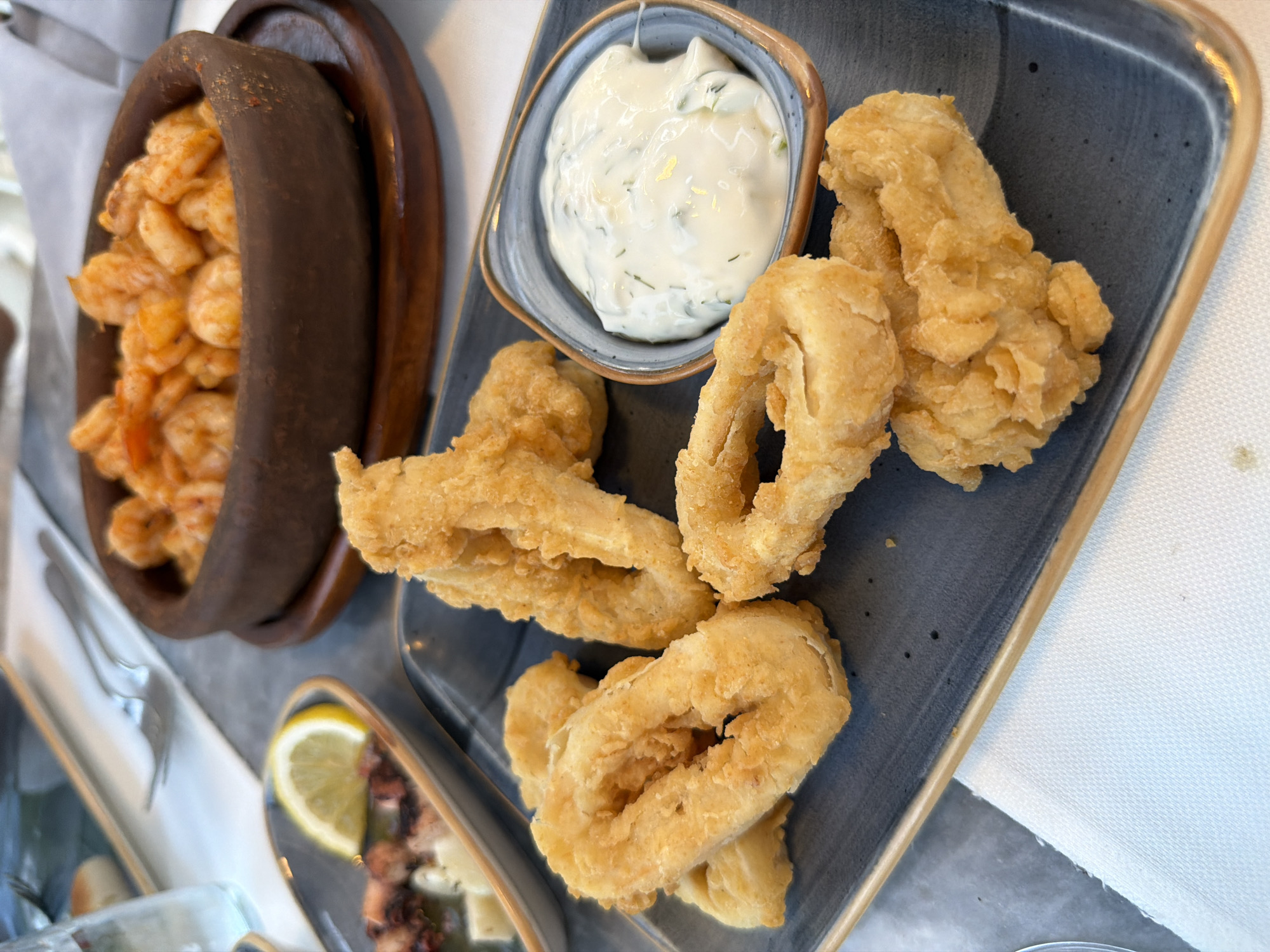 A close-up, eye-level shot captures a meal with several seafood dishes on a table in what appears to be a restaurant setting in Kemer, Turkey. In the foreground, a dark blue rectangular plate holds a generous serving of golden-brown, battered calamari rings. A small, angled blue bowl filled with a creamy white dipping sauce, speckled with green herbs, sits to the right of the calamari. Behind the calamari, a small oval dish contains pieces of what looks like marinated octopus with a slice of lemon. Further back, a rustic brown earthenware dish is filled with cooked shrimp in a reddish sauce. The table is set with white napkins and silverware, suggesting a dining experience. The lighting is natural and soft, highlighting the textures and colors of the food.