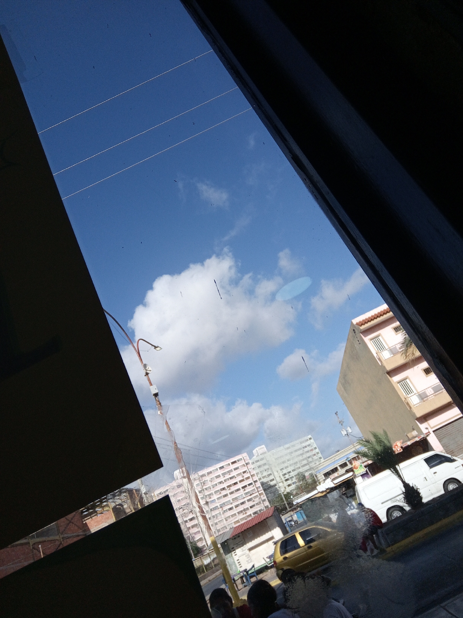 The image shows a view from inside a building, likely a storefront or restaurant, looking out onto a street in Juan Griego, Venezuela. The view is partially obstructed by the window frame and interior elements. The sky is blue with scattered clouds. Buildings of varying heights and colors are visible in the background, including a tall pink apartment building, a lighter grey building, and a shorter tan building. On the street, there is a yellow car, and a white van. Two people appear to be interacting by the curb, appearing blurred. Streetlights are visible along the roadway. Reflections are apparent on the window surface.