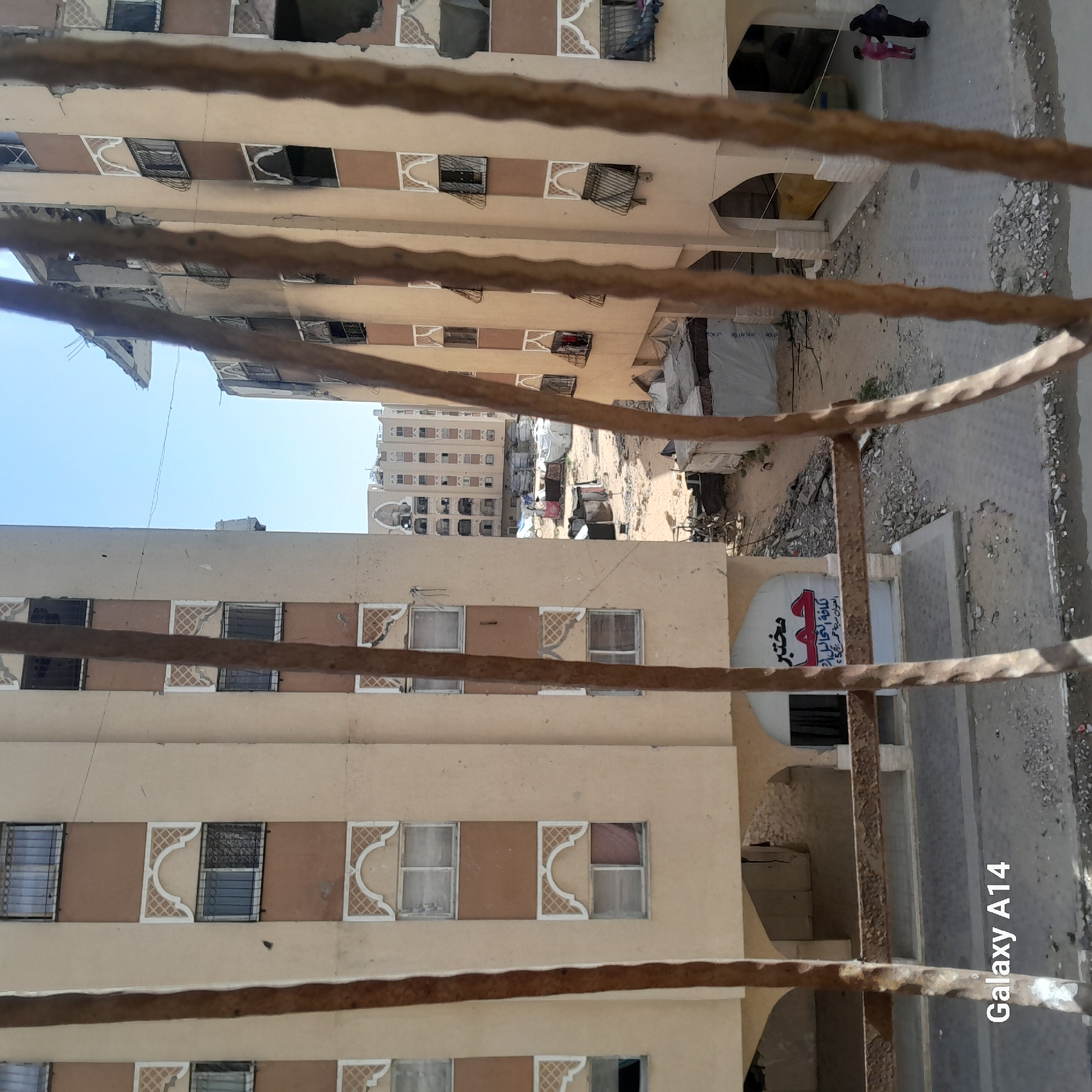 Here is a detailed description of the image:
**Scene Overview:**
The image presents an eye-level view from a balcony or window, looking out over a city, likely taken in an urban area in Unknown, Unknown. The perspective is partially obstructed by rusty metal bars, suggesting the photograph was taken from inside a building.
**Environment:**
The cityscape is dominated by residential buildings of similar design and color schemes. The buildings feature multiple stories with repetitive patterns of windows and architectural details. Some buildings show signs of damage, possibly from conflict or natural disaster. There is a significant amount of debris and rubble visible, indicating a post-conflict environment.
**Objects and Details:**
* Buildings: The buildings are multi-story and have similar architectural elements like decorative window frames and beige-colored facades.
* Debris: Piles of rubble and debris are scattered throughout the scene, especially in the areas between the buildings.
* Signage: A white sign with writing in Arabic is visible.
* Balcony/Window: The image is taken through or from a balcony/window featuring rusty metal bars.
* Garbage/Refuse: Appears to be a build-up of refuse and trash in between and next to buildings.
**People:**
There are two people in a courtyard, they seem to be moving a long, white object, possibly wrapped in plastic.
**Overall Impression:**
The image portrays a damaged urban environment with signs of conflict or disaster. The combination of the damaged buildings, the presence of debris, and the visible signage provide a sense of location and context.