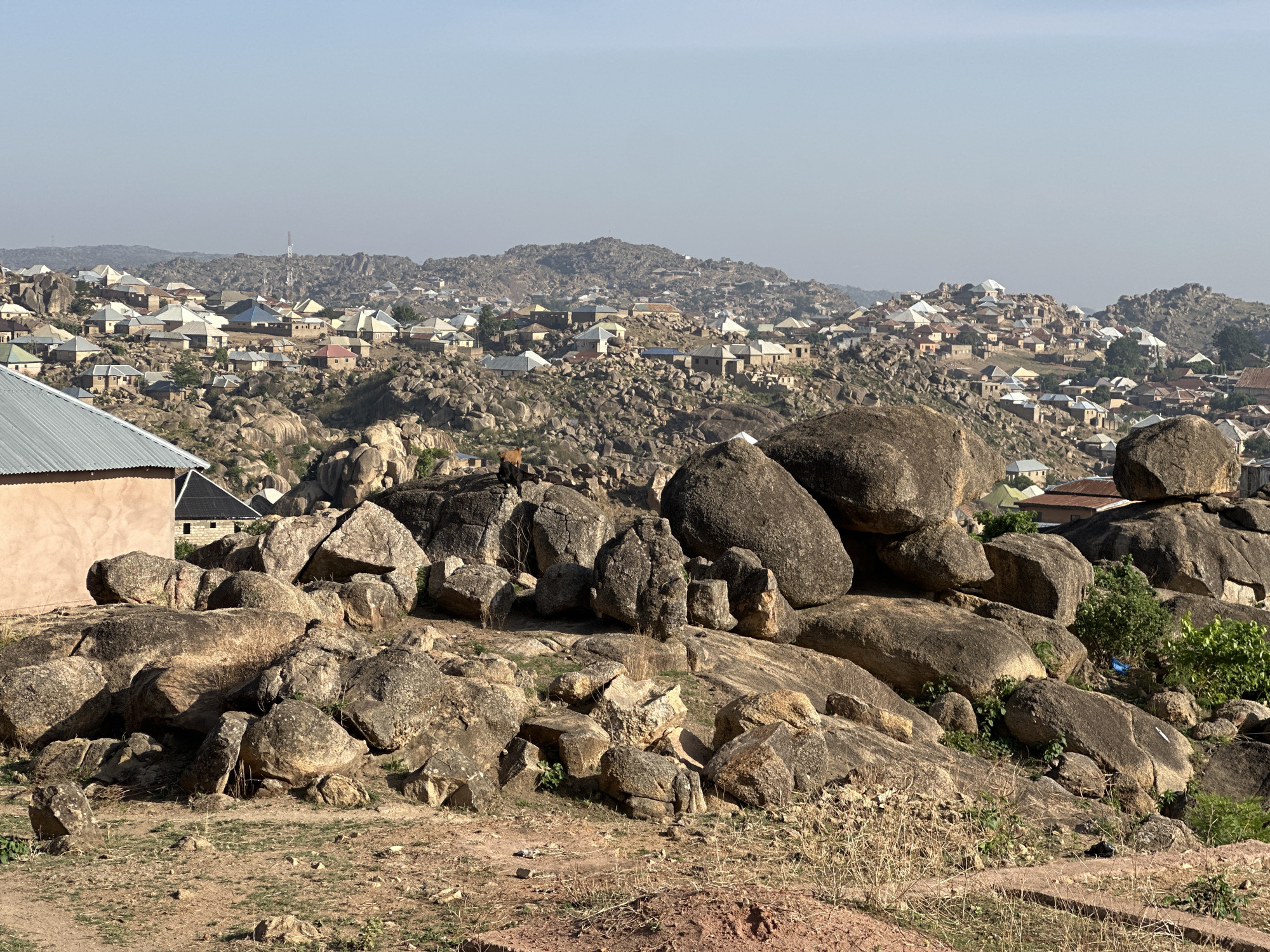 The image shows a landscape in Jos, Nigeria, characterized by a settlement built among large granite rock formations. In the foreground, there's a cluster of big, rounded brown rocks with some vegetation. The ground is dry and reddish-brown. Behind these rocks lies a densely populated area with houses scattered across the hills and rocks. The houses have tin or corrugated metal roofs, mostly gray. In the distance, there are more rocky hills with houses, and a communication tower is also visible. The sky is pale blue and hazy, suggesting a dry climate. The overall scene captures the unique way of life in Jos, where people have adapted to living within a rocky environment.