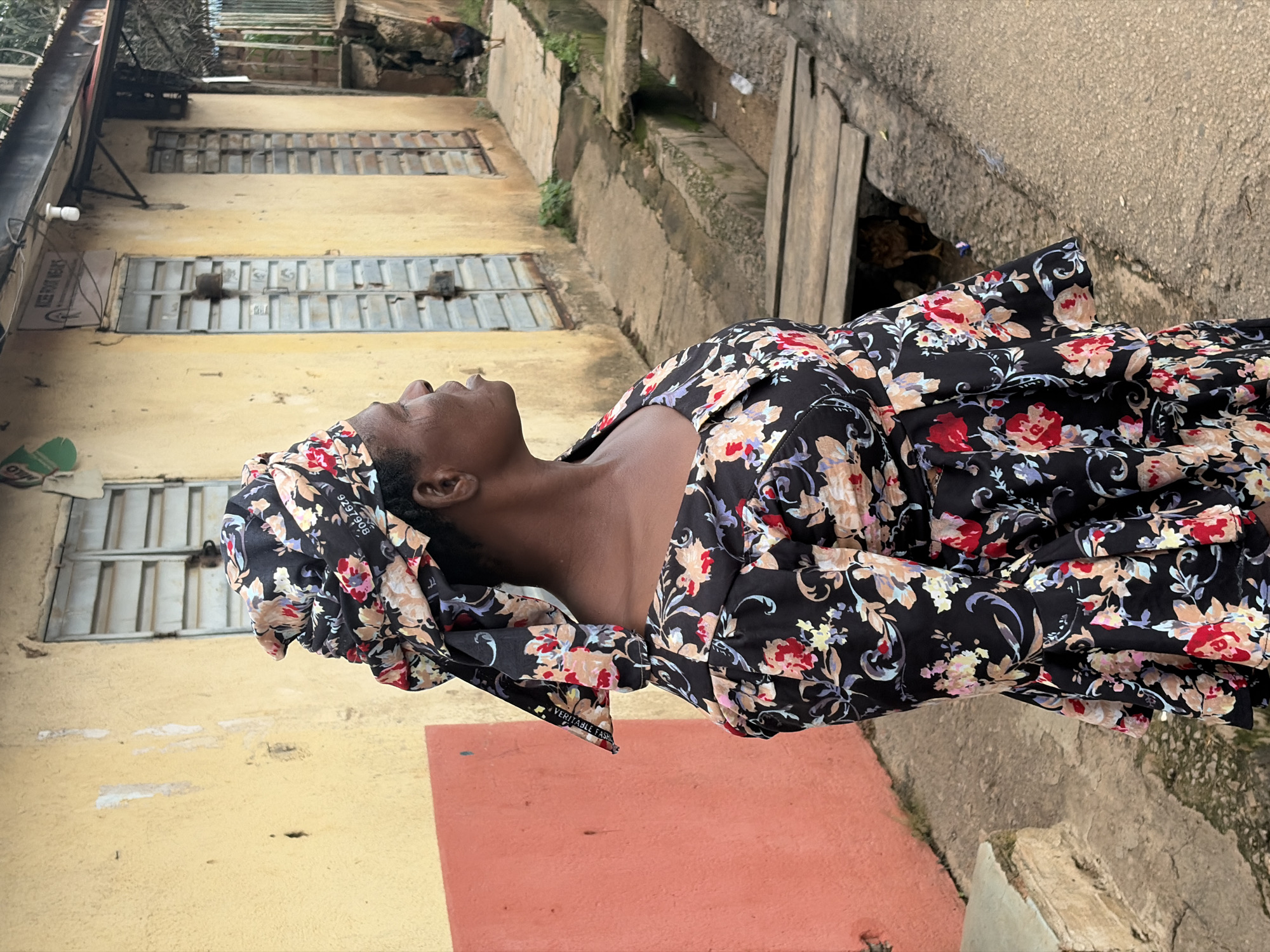 In the city of Jos, Nigeria, a woman stands on a dirt road facing away from the camera, her head turned to the right in profile. She wears a black floral-patterned headwrap and a matching dress with puffy sleeves and a ruffled hem. Behind her, a pale yellow building with faded blue shutters stands on the left, while a darker building with a corrugated metal roof is visible on the right. A narrow, open concrete drainage ditch runs along the side of the road, spanned by a makeshift wooden bridge. A rooster stands in the background on a paved path in front of the darker building. The scene appears to be an outdoor market or residential street.