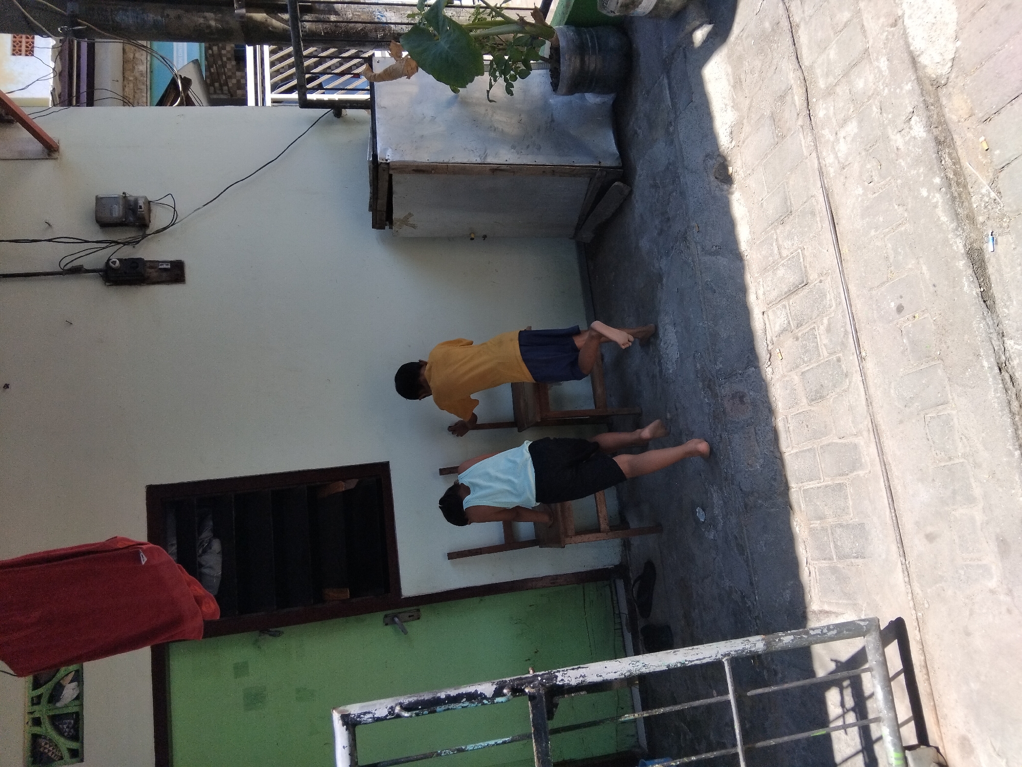 Two boys are near a house in Kepuh Kiriman, Indonesia. The boy on the left, wearing a light blue tank top and black shorts, is leaning on a wooden chair. The boy on the right, in a yellow t-shirt and blue shorts, is standing next to another wooden chair. A red garment hangs from a line above a green door. Electrical meters and wires are visible on the wall of the house. A metal cabinet and a potted plant are also present. The ground is paved with concrete blocks.