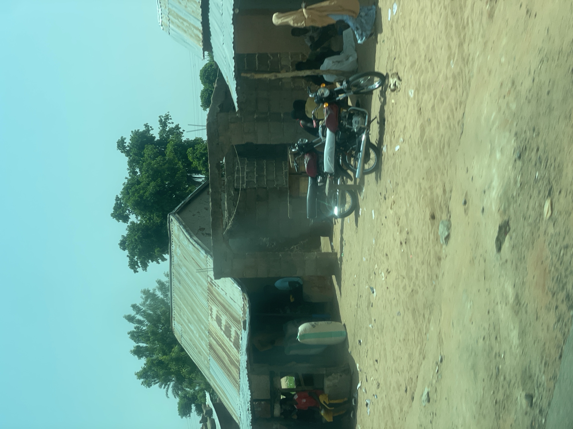 The image depicts a scene in Zongo Mari, Nigeria, featuring a dusty, outdoor environment with several buildings and people. In the foreground, a sandy area is littered with debris. Two motorcycles are parked prominently. Behind the motorcycles, a group of people are gathered, some sitting on the ground and others standing. To the left, a building with a corrugated metal roof has an open front where a person in a red shirt and yellow pants is seated. Another person in a blue shirt is visible inside. A large, striped sack is placed near the entrance of this building. Further to the right, another building with a similar metal roof is seen, with more people gathered in its shade. The sky is a light blue with some haze. The overall impression is of a community gathering or marketplace in a rural or semi-rural setting.