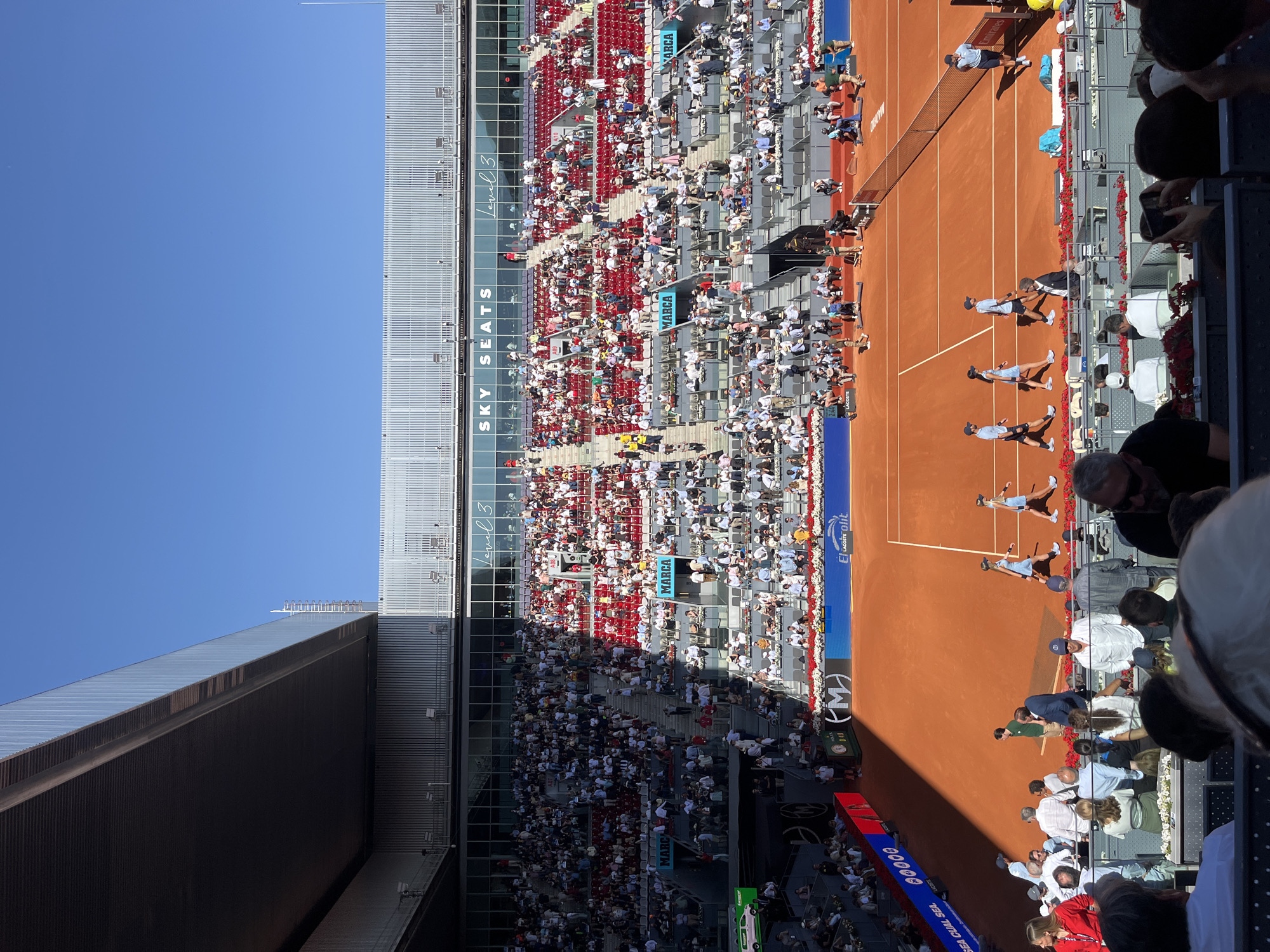 This image captures a wide view of a professional tennis match underway on a sunny day in Madrid, Spain. The scene is set in a large outdoor stadium, featuring a distinctive orange-red clay court.
On the court, several male tennis players are visible, some actively playing or preparing for a serve/return, and others potentially officials or coaches. The court is clearly marked with white lines and features a net across the middle.
The stadium grandstands are filled with numerous spectators, particularly in the sections closest to the court. The seating areas are a mix of red and grey seats, with some sections appearing more crowded than others. Above the main spectator areas, a section labeled "SKY SEATS" is visible, along with multiple "Level 3" markers, indicating different tiers of seating or stadium levels. The architecture of the stadium includes large, modern-looking structures with corrugated panels and glass windows.
The weather appears clear and bright, with a vibrant blue sky dominating the upper left portion of the image. Strong sunlight casts distinct shadows across parts of the court and stands, suggesting it is midday or early afternoon. The perspective is from an elevated position in the stands, looking down and across the court.
Visible text and logos include: "SKY SEATS", "Level 3" (twice), "MARCA" (a Spanish newspaper, seen multiple times as an advertisement), "EL LACOSTE" (likely associated with the Lacoste brand), "SEA CUAL SEA", "00000", and an "(M)" logo. Some spectators are seen holding phones, potentially recording the match.
