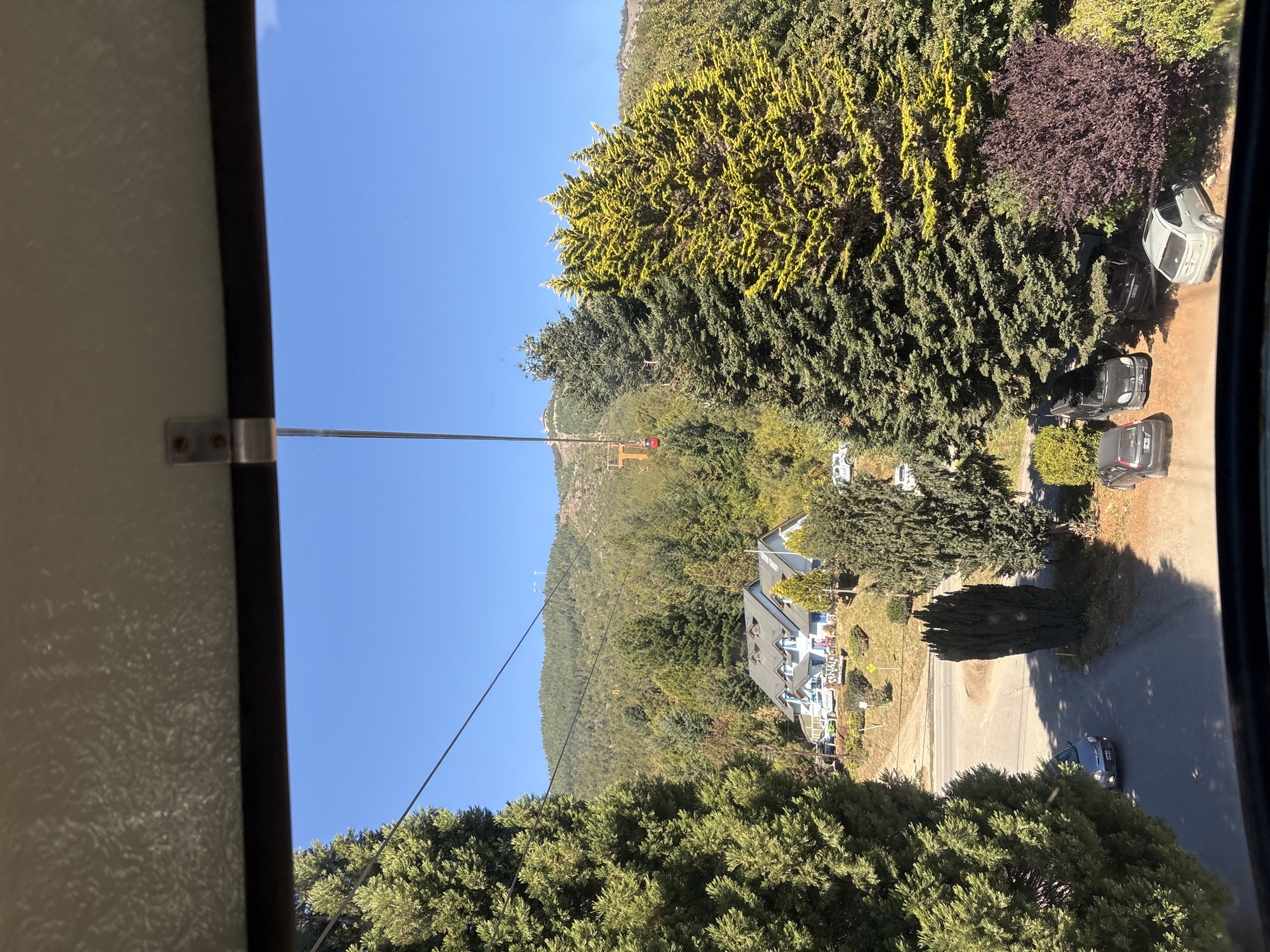 The image depicts an elevated view of a mountainous, residential landscape in San Carlos de Bariloche, Argentina, captured from inside a gondola cabin. The sky is clear and brilliant blue, dominating the upper frame. Below the sky, a verdant mountain range covered densely with coniferous trees, some exhibiting a yellowish-green hue, extends into the background.
A prominent light-blue, multi-story building with white trim, a dark grey roof, and multiple dormer windows is situated centrally on a gently sloping hill. Surrounding the building are various trees, shrubs, and a winding paved road. Multiple vehicles are visible; a silver hatchback is parked on the paved road in the lower-left, and several others are parked on unpaved surfaces: a black van, a dark sedan, another dark vehicle, and a light-colored SUV.
A gondola cable diagonally traverses the scene from the lower-left towards the upper-right. A single red gondola cabin is visible mid-air in the mid-ground, ascending towards a distant mountain peak where a chairlift tower and other smaller structures are discernible. No individuals are explicitly present in the frame, but the infrastructure and vehicles indicate human presence and activity within the environment.