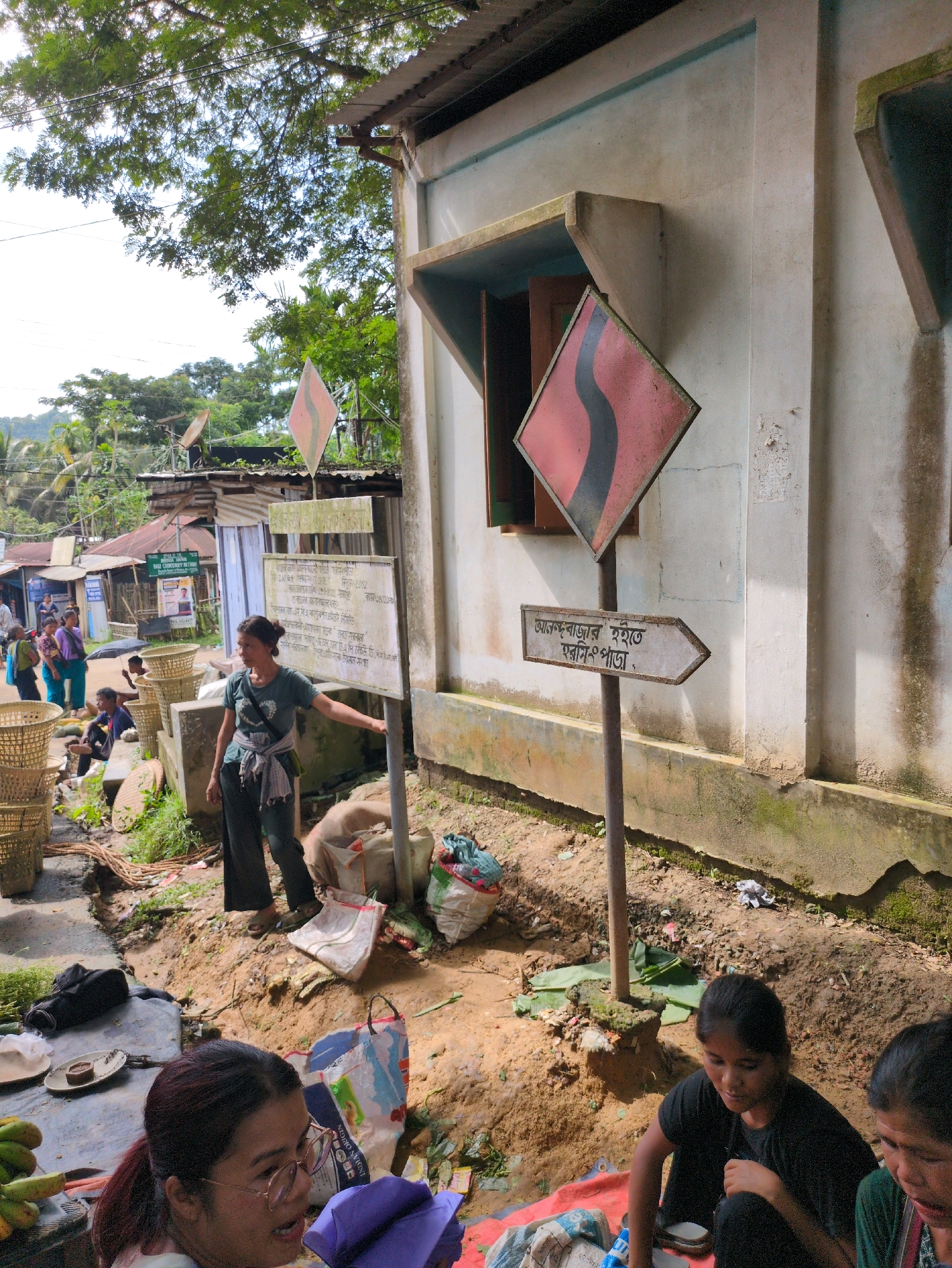 In Borchora, India, a woman stands outdoors, gesturing towards a directional sign. The sign is composed of a diamond-shaped warning sign above a rectangular directional sign. The directional sign, with text in a foreign script, points to the right. The woman, dressed in a dark t-shirt and pants, has her hair tied back and is wearing a scarf around her waist. She is standing on a dirt path, with a building behind her. The building has a light-colored facade and a metal roof. In the foreground, various items are laid out on the ground, including baskets, bags, and produce, suggesting a marketplace or roadside stall. Other people are visible in the background, some standing and others sitting. The environment appears to be rural or semi-urban, with trees and smaller structures visible.