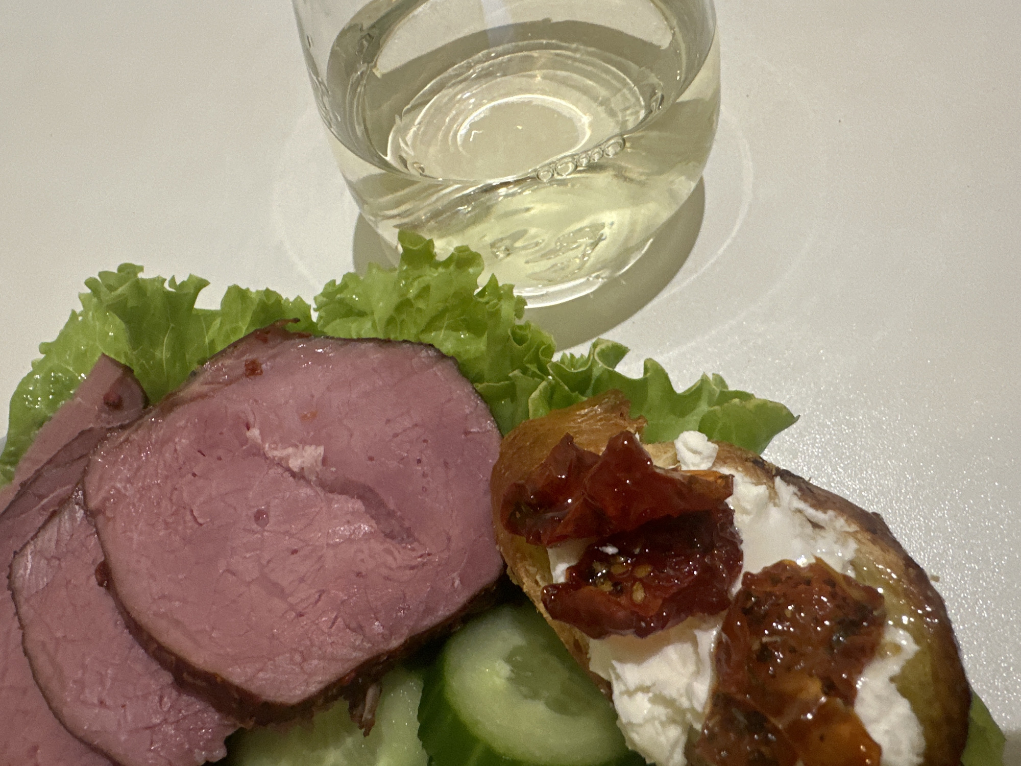 A close-up shot depicts a glass of light-colored liquid, possibly white wine, next to a plate of food. The food includes sliced meat, lettuce, sliced cucumbers, and bread topped with white cheese and what appears to be a tomato-based topping. The environment suggests an indoor setting, possibly a restaurant or home. The location mentioned is Chaikovsky, Russia. There are no people visible in the image.