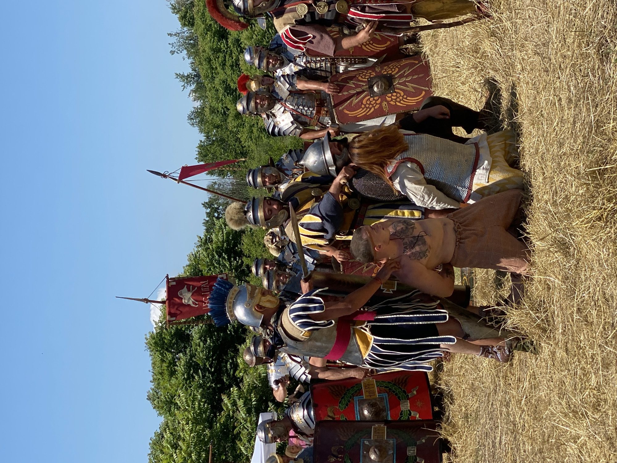 The image depicts a historical reenactment scene taking place outdoors in Gigen, Bulgaria, under a clear blue sky with bright sunlight. The ground is covered with dry, light-colored straw or hay. In the background, dense green foliage (trees and bushes) is visible.
At the center of the foreground, two individuals are kneeling on the dry ground. The person on the left is a man, bare-chested, revealing tattoos on his upper left shoulder and back, and wearing light-colored, possibly linen, lower garments. The person on the right is a woman with long reddish-brown hair, wearing a light-colored tunic and a light-colored, possibly padded or scale-like, upper garment.
Surrounding the kneeling individuals are numerous participants dressed as Roman soldiers. A prominent soldier standing over the kneeling man wears a silver Roman helmet with a blue and white crest, segmented plate armor (lorica segmentata), a red sash, and striped pteruges (skirt-like strips) in blue and white. He extends his right hand towards the kneeling man. Another soldier, standing over the kneeling woman, wears a silver helmet and chainmail (lorica hamata) over a tunic, and points downwards with his right hand.
Other Roman soldiers are positioned in the midground and background. They are equipped with various forms of Roman armor including lorica segmentata and lorica hamata, helmets (some with red plumes), and tunics in colors such as red, yellow, and white. Many soldiers carry large, rectangular scutum shields, predominantly red with gold-colored winged or wreath designs and central metallic bosses.
Two standards are visible: one red rectangular banner on a pole in the left background features a white animal (possibly a wolf or lion) and the inscription "LEG ITAL". Another red, triangular pennant is seen on a pole in the right midground. A white canopy or tent structure is partially visible in the far left background. The overall scene suggests a staged event, possibly a depiction of capture, judgment, or a ceremonial gathering.