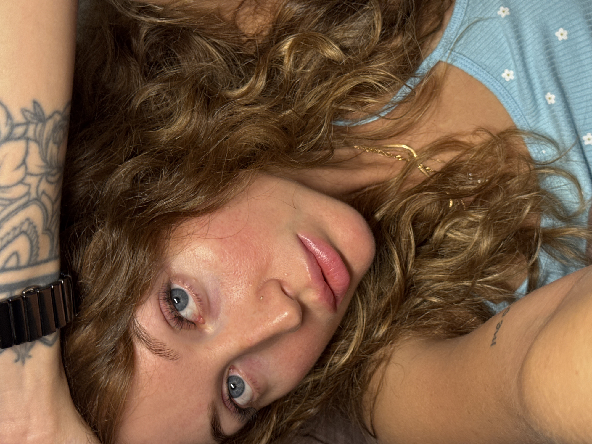 A young woman with voluminous, wavy, light brown hair frames her face as she lies down, looking directly at the viewer with an intense, yet calm gaze from her light blue eyes. Her skin has a natural, slightly flushed appearance, and her full, naturally pink lips are softly parted. A small, subtle stud piercing is visible on her right nostril.
On her left forearm (viewer's right), a large, intricate tattoo featuring floral or botanical patterns is clearly visible in a grayscale style. A dark-colored watch with a metallic link band is worn on the same wrist. She is wearing a light blue top adorned with small, white daisy-like patterns, partially visible on her upper body. A thin gold chain necklace can be seen around her neck, peeking through her hair. On her right upper arm (viewer's left), a small text tattoo is present, with the word "me" discernible.
The background is indistinct, largely obscured by her hair and body, suggesting an intimate indoor setting, possibly on a bed or floor. The lighting is soft and even, highlighting her features. The woman is posing for a close-up photograph, conveying a direct and engaging demeanor. While the image is set in Chaikovsky, Russia, no specific geographical markers are visible within the scene itself.