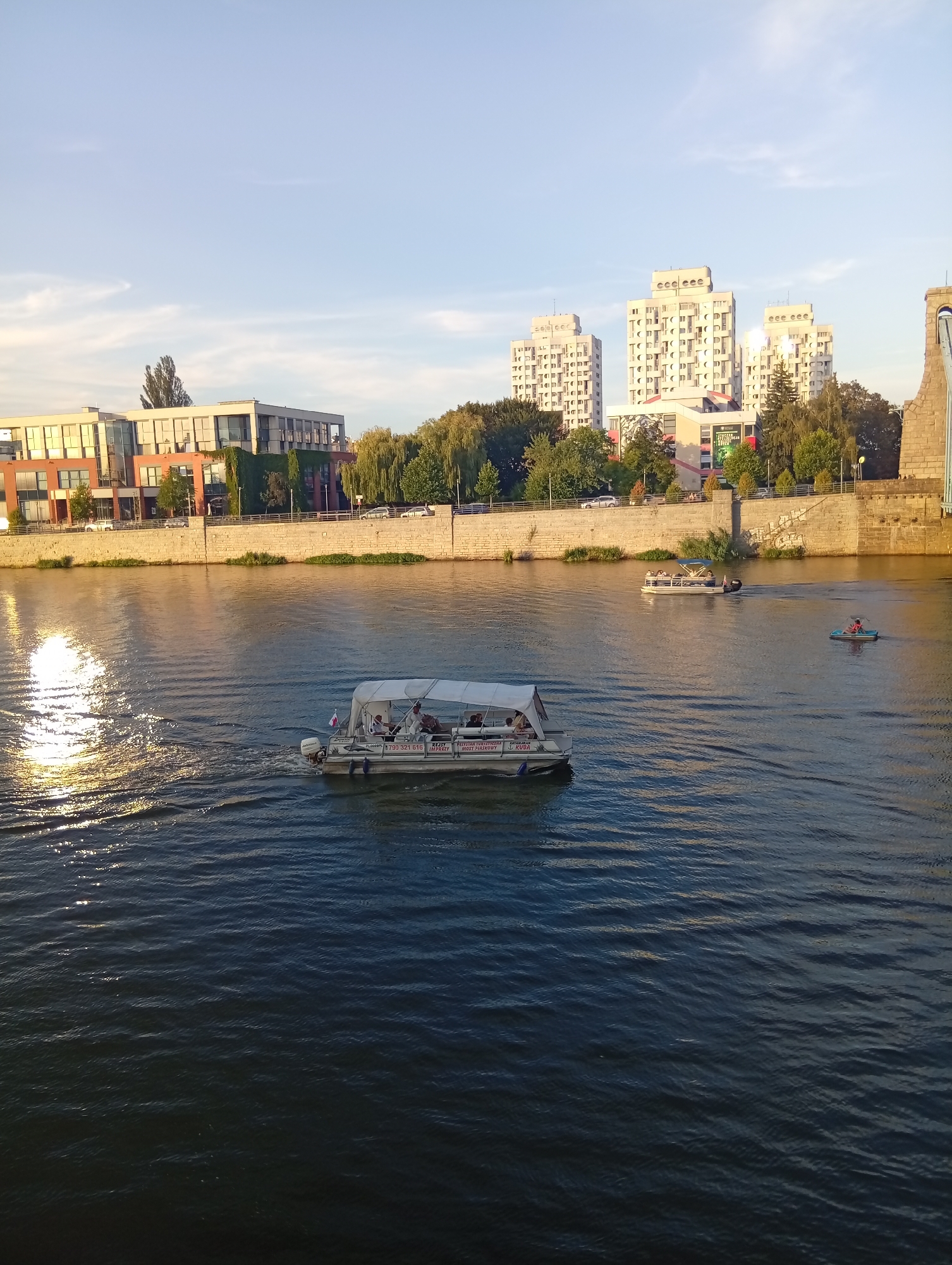 In Wrocław, Poland, a river flows through a cityscape bathed in late afternoon sunlight. The water's surface is rippled, reflecting the warm glow of the setting sun on the left side of the frame. Several boats navigate the river: in the foreground, a pontoon boat with a canopy carries several passengers; further in the distance, another pontoon boat is visible, and a person in a small blue boat is seen on the right.
Along the riverbank, a stone embankment supports a row of buildings. On the left, a modern brick building with large windows is prominent. Behind it, a line of trees separates it from more distant, contemporary white high-rise apartment buildings. To the right of the apartment buildings, lush green trees line the bank. The sky above is a clear blue, with a few wispy white clouds. A portion of a large bridge structure is visible on the far right edge of the image. The scene captures a tranquil moment of urban life by the water.