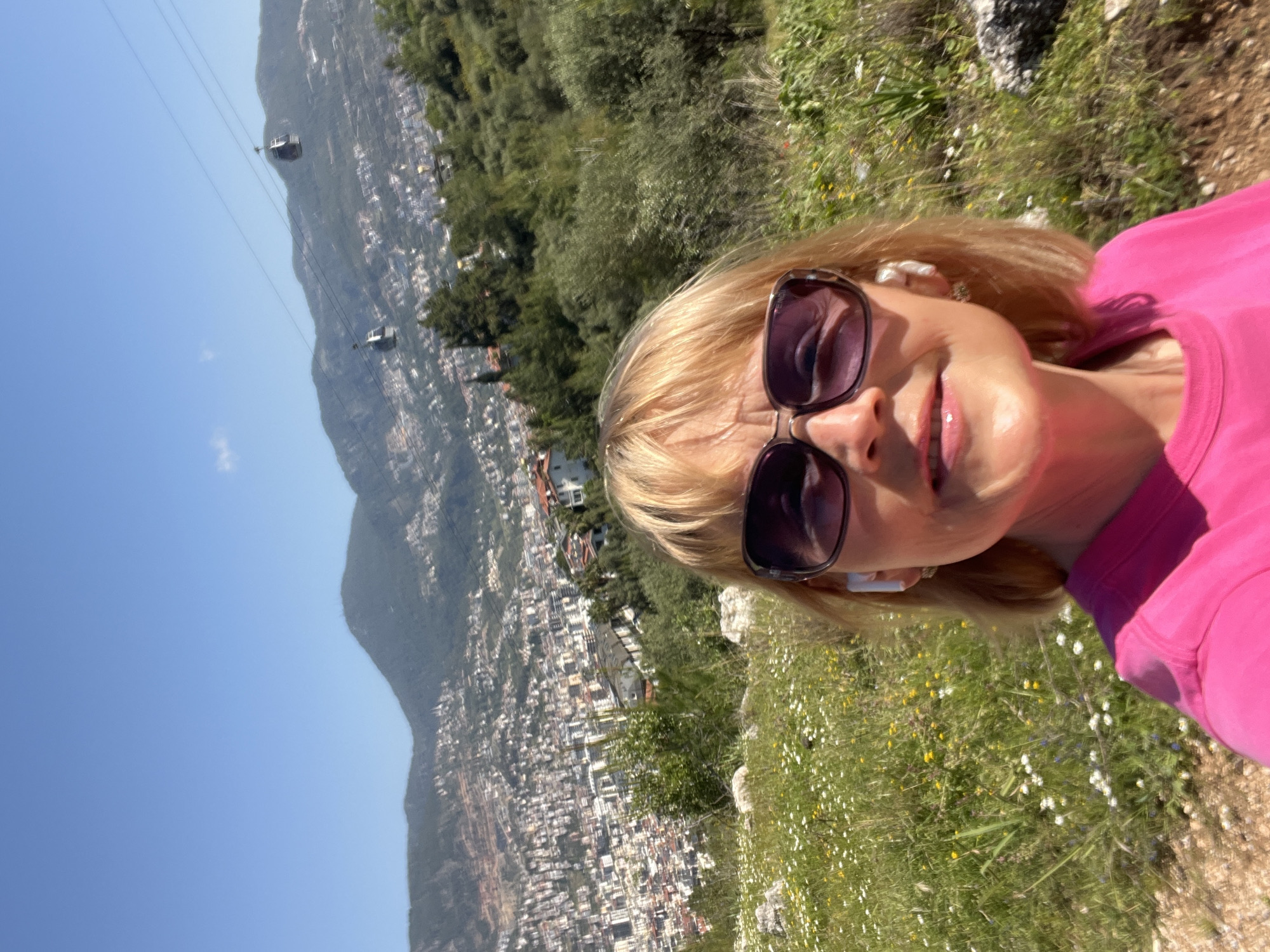 The image is a bright, sunlit selfie-style shot of a blonde woman smiling into the camera. She is wearing large, dark sunglasses, with the brand "Fendi" visible on the arm, and a vibrant pink top. White earbuds are visible in her ears.
The background presents a sweeping panoramic view from an elevated position. Below, a bustling city or town spreads across the slopes of a mountain or large hill, characterized by numerous white and light-colored buildings nestled amidst green foliage. In the mid-distance, two cable cars are suspended on lines, moving across the clear blue sky, with more mountainous terrain visible beyond the urban development. The immediate foreground around the woman shows natural ground covered with green grass and small white and yellow wildflowers.
The time of day is clearly daytime, with bright sunshine and a cloudless blue sky, suggesting pleasant weather. The woman's smile indicates enjoyment. The scene's elements—mountains, city on slopes, cable cars, and coastal-like vegetation—are consistent with the provided location context of Alanya, Turkey, suggesting the image was taken from a viewpoint overlooking the city.