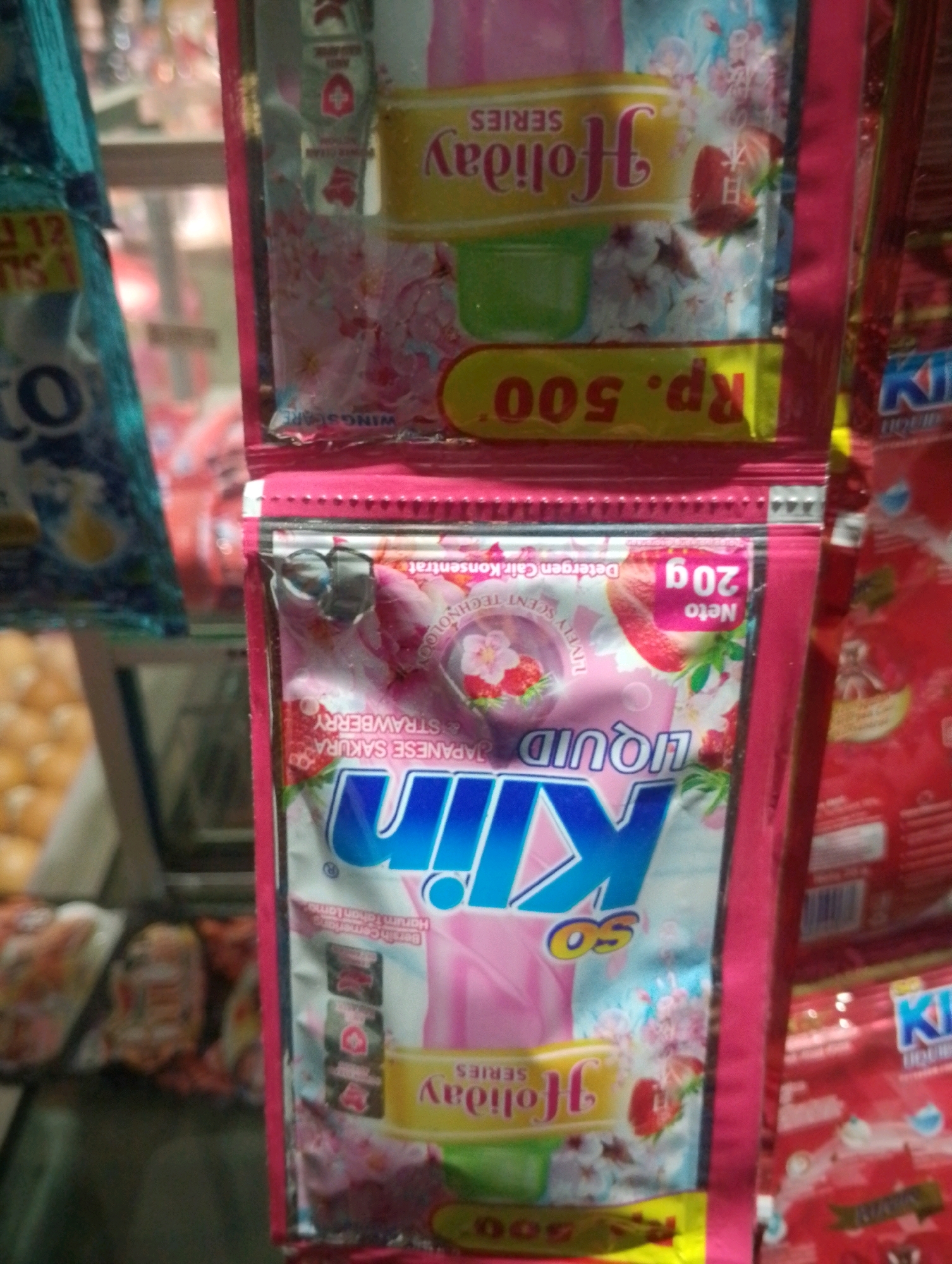 The image shows a close-up of several brightly colored product packages, likely detergent or soap, displayed vertically. The dominant colors are pink, blue, and yellow. In the foreground, there are two pink packages of "LIQUID KIM" detergent. One package features a prominent blue "KIM" logo with a circle inside the "i," and text including "JAPANESE SAKURA" and "STRAWBERRY." Another package, also pink, is partially visible to the left and has "SERIES" and "HOLIDAY" text in yellow and pink. The bottom portion of the image shows more packaging, including what appears to be a price tag with "Rp. 500". The background is blurred, but suggests a retail environment, possibly a market stall or shop shelf, with other products and shelves visible. The lighting is artificial and casts a slight glare on the packaging. The overall impression is of a retail display of personal care products.