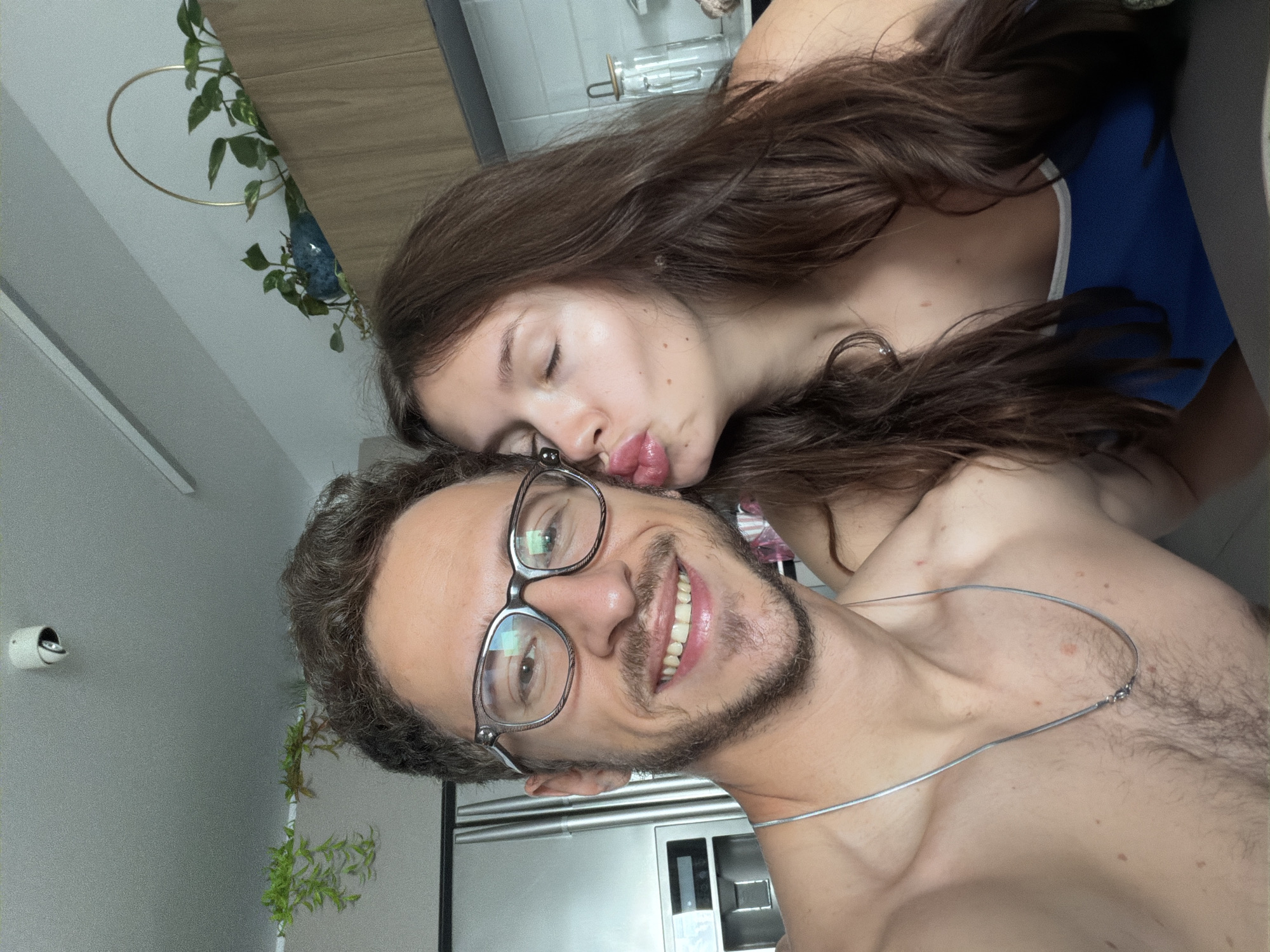 A man and a woman are captured in a close-up, diagonally oriented selfie, suggesting a casual and affectionate moment. The man, in the foreground, smiles broadly at the camera, his teeth visible. He wears clear-framed glasses with reflective lenses and has short, dark brown curly hair and a beard. He is shirtless, displaying his chest and shoulders, and a thin silver necklace.
Behind and slightly to his left, the woman rests her head, with her eyes closed and lips puckered into a playful "kiss" expression. She has long, wavy dark brown hair. A blue top with a white trim is visible on her.
The background indicates an indoor domestic setting, likely a modern apartment in Tel-Aviv, Israel. A white, cylindrical security camera is mounted on a light gray wall in the upper left. Green foliage from potted or hanging plants is visible in various parts of the background, including one plant arranged within a golden hoop. Parts of a modern kitchen are discernible behind the man, featuring what appears to be a stainless steel refrigerator or built-in appliance and a sink area. A clear glass container is also visible behind the woman's head. The overall impression is one of happiness and intimacy between the couple.