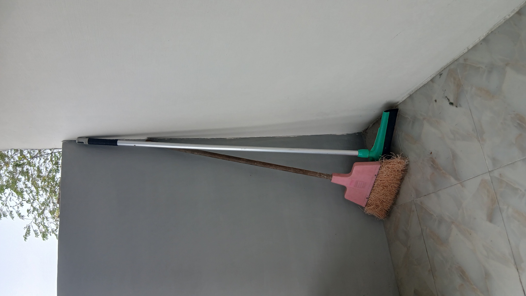In the city of Klambir Lima Kebun, Indonesia, two brooms are leaning against the corner of a light-colored wall. One broom has a pink plastic head and a natural fiber brush, with a wooden handle. The other broom is green with a black rubber squeegee and a metal handle. Both brooms are standing upright on a tiled floor, which appears to be outdoors, given the glimpse of trees and sky at the top of the frame. The environment suggests a simple, utilitarian setting, possibly a porch or patio area.