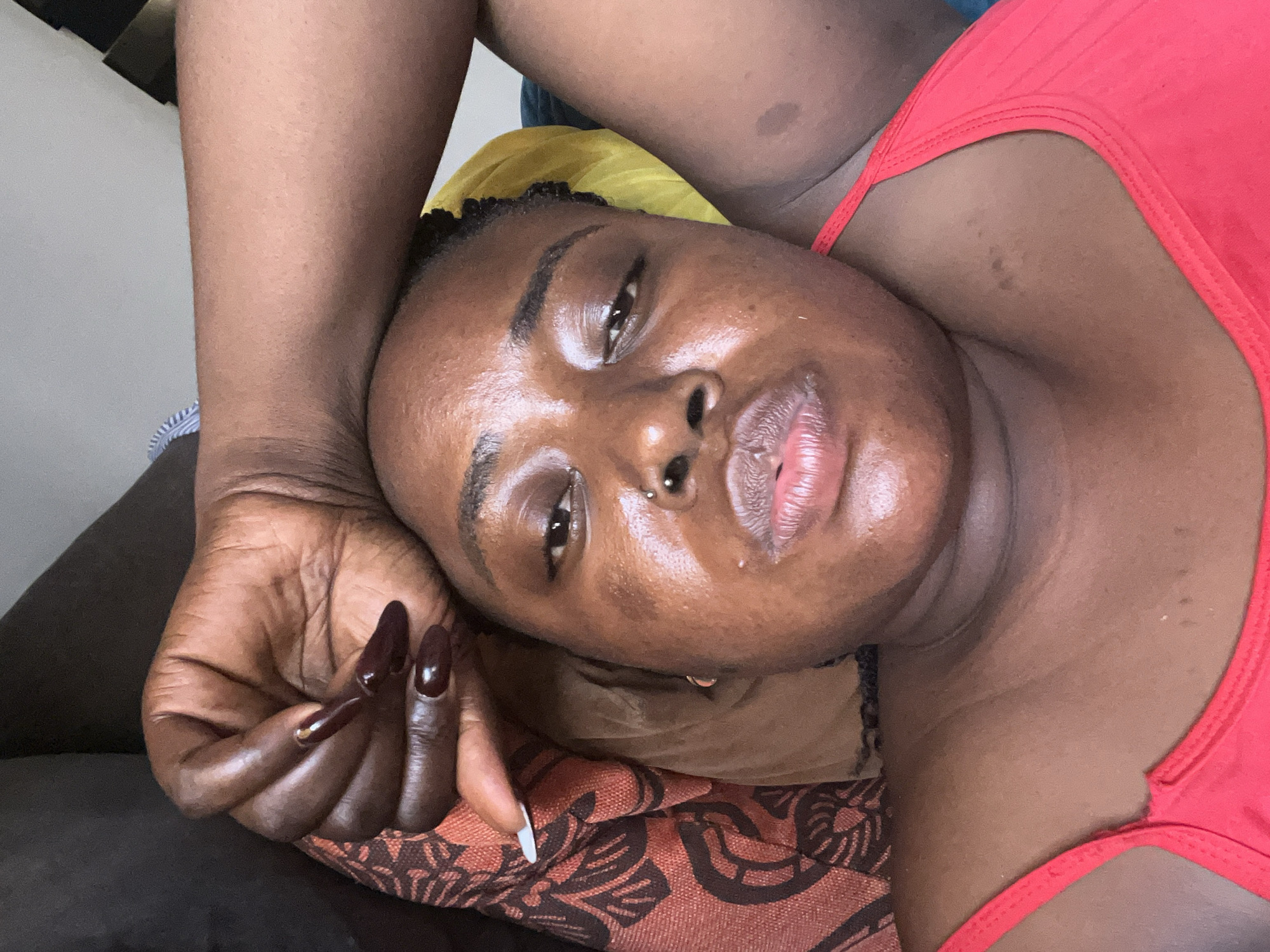The image is a close-up selfie of a dark-skinned woman lying down, with her left arm raised and resting on her forehead. She is wearing a red top. Her facial features are prominent, including her eyes, nose with a septum piercing, and lips. Her right hand, with long, dark, almond-shaped nails, is also visible near her face. The background is slightly blurred, showing a patterned fabric to her left and a yellow object behind her head. The lighting suggests an indoor setting. There is no visible indication of the city of Abuja, Nigeria.