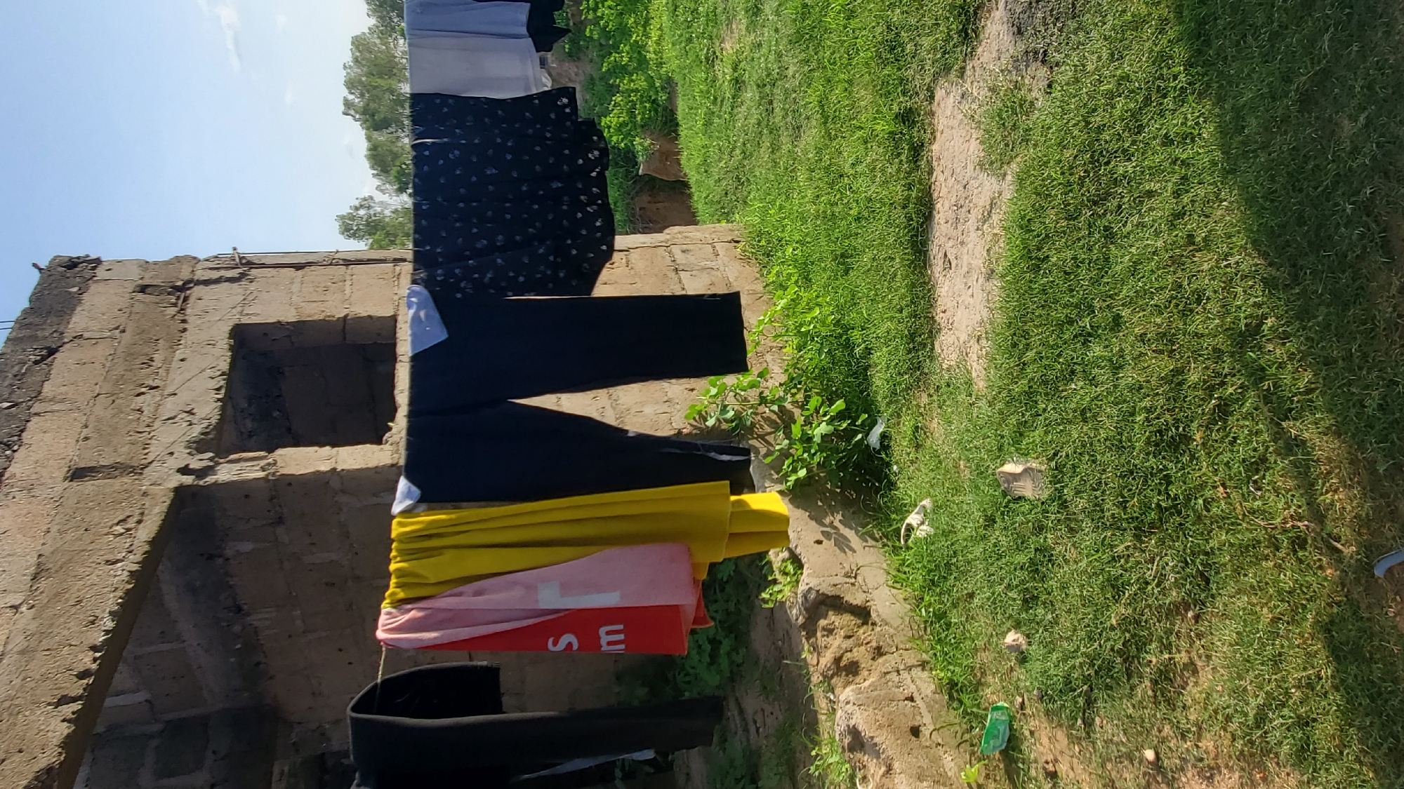 The image depicts an outdoor scene in Jalingo, Nigeria, featuring a clothesline strung with various garments. The clothesline is suspended in front of an unfinished, brick structure, characterized by exposed blocks and irregular openings. Lush green grass covers the ground, interspersed with dirt patches and some scattered debris, including what appear to be plastic bags and possibly a discarded water bottle. Several items of clothing are hanging on the line, including a red garment with white lettering, a pink garment, a yellow garment, dark-colored pants, a black garment with a white floral pattern, and a light-colored garment, likely a shirt. The sky above is a clear blue, suggesting fair weather. The overall impression is one of a residential area in an early stage of construction, with everyday activities like drying laundry taking place.