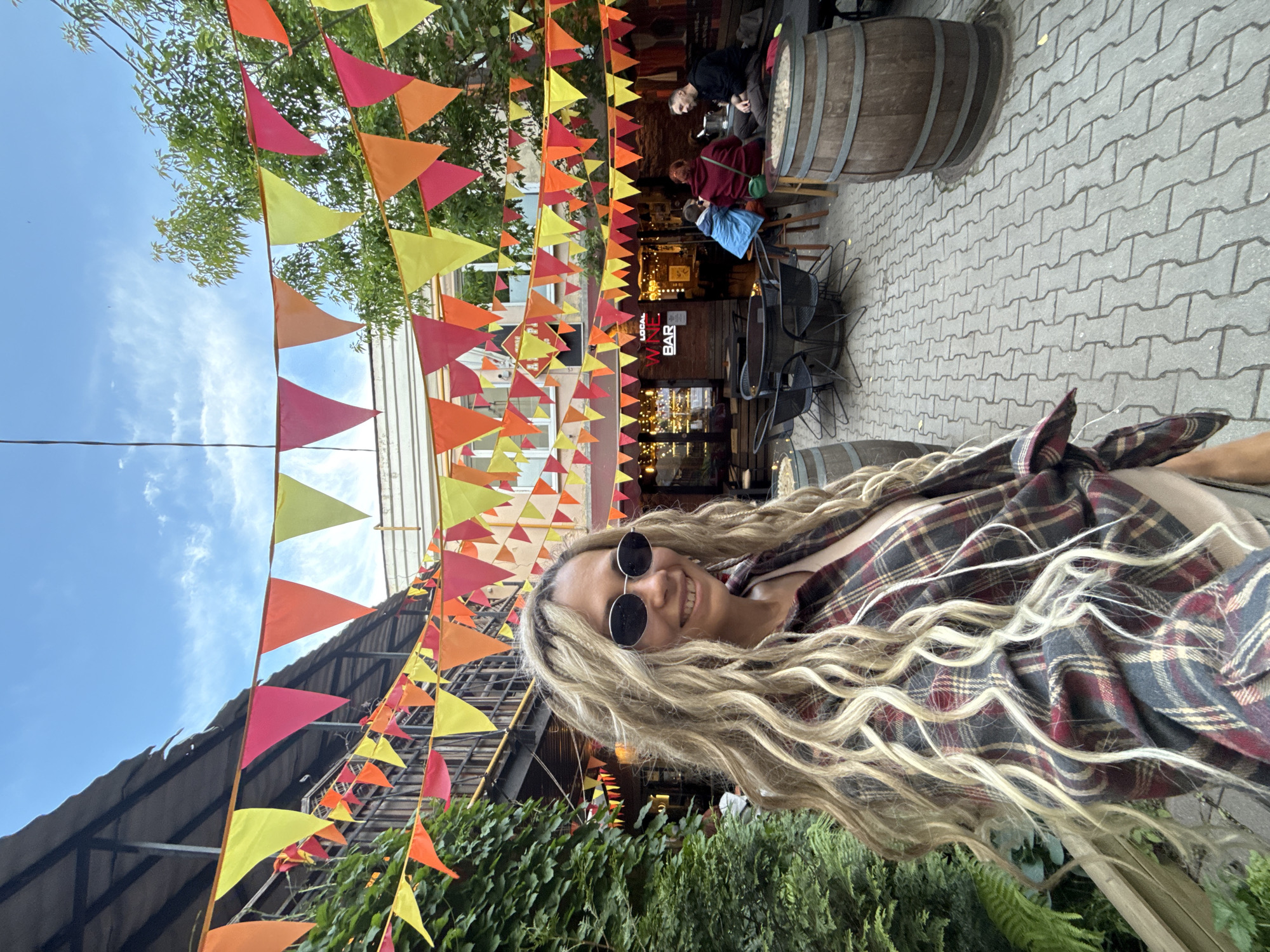 This vertically-oriented selfie, rotated 90 degrees clockwise, captures a woman smiling broadly in an outdoor, festive setting. The main subject, positioned on the right side of the frame, has long, wavy blonde hair and wears dark, round sunglasses and a plaid shirt in shades of red, grey, and cream. Her cheerful expression suggests enjoyment.
The scene is an outdoor area, possibly a street, market, or restaurant patio, adorned with numerous colorful triangular pennant flags strung overhead. These flags are in vibrant hues of yellow, orange, pink, and red, creating a festive atmosphere.
In the upper left background, several people are seated at outdoor tables outside what appears to be a bar. One man with dark hair is visible from the side, while others in red and blue attire are partially obscured. Large wooden barrels with metal bands are used as décor or tables around this area. The ground is paved with grey interlocking concrete blocks.
Visible text on a sign above the bar entrance reads "LOCAL WINE BAR," though partially covered by the flags. Green foliage, resembling shrubs or ivy, adds natural elements to the foreground and background. The clear blue sky with some wispy clouds indicates daytime and good weather. The overall impression is that of a lively, casual outdoor gathering in Rusanovo, Russia.