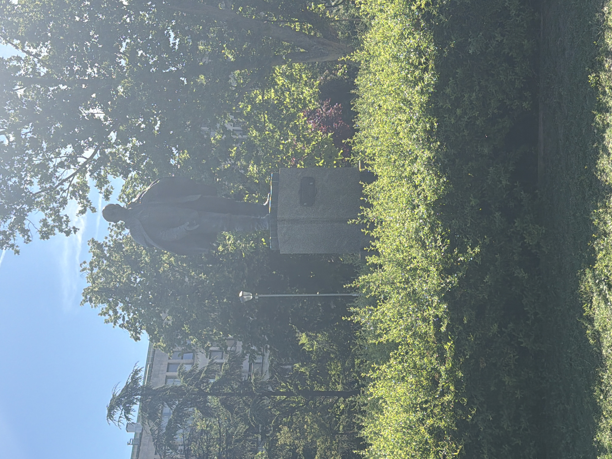 The image depicts a statue of Jovan Cvijic located in Belgrade, Serbia. The statue is positioned on a stone pedestal and surrounded by trees and a neatly trimmed hedge. A streetlight is partially visible behind the statue. In the background, a building can be seen. The weather appears sunny.