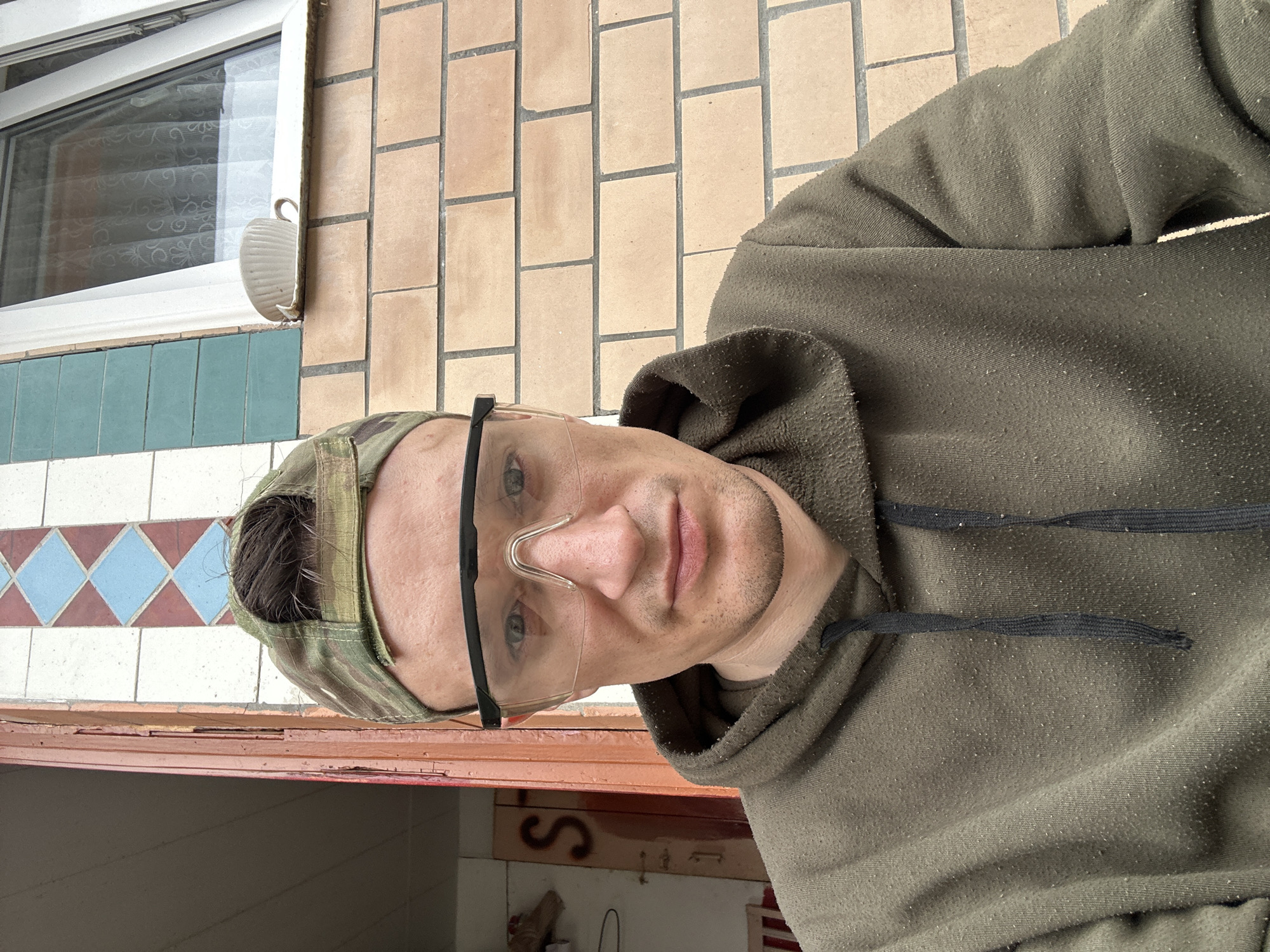 In Makiv, Ukraine, a man wearing safety glasses and a camouflage baseball cap is seated outdoors. He is dressed in a dark green hooded sweatshirt. Behind him, a brick wall forms the backdrop, with a window visible above a row of bricks. To his left, a wooden door frame is partially visible, with some kind of writing in red on the door. The man appears to be in the process of taking a selfie.