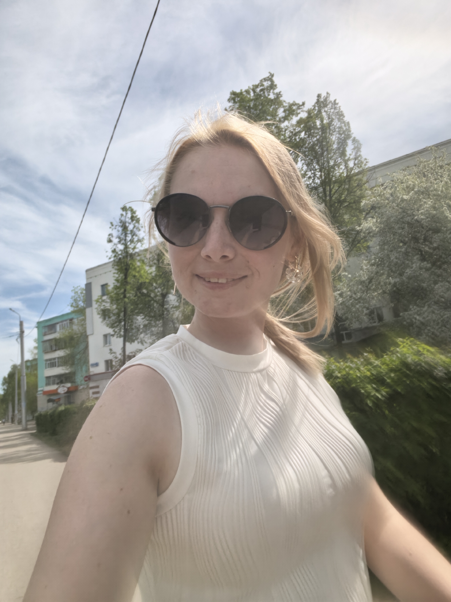 A fair-skinned woman with blonde hair, wearing sunglasses and a white sleeveless top, is taking a selfie. The woman is smiling slightly, and her hair is pulled back in a ponytail. Behind her is a street with buildings and trees. A powerline is visible in the upper left corner. The buildings appear to be apartment complexes with at least one featuring a green façade. The location is likely in Sterlitamak, Russia, suggested by the architecture and environment. Some of the trees are flowering, indicating springtime. The image is somewhat blurred, implying motion during capture.