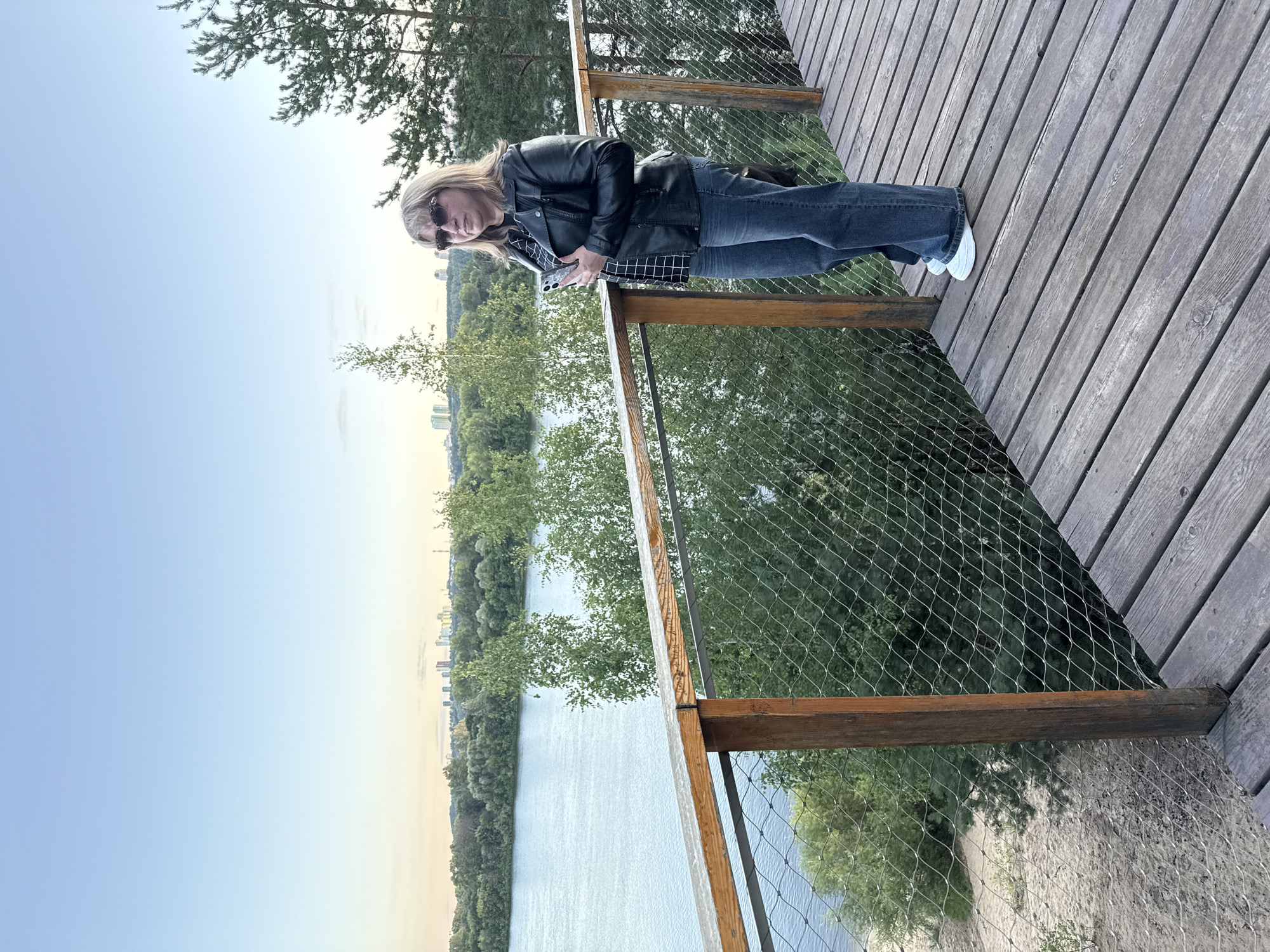 A woman stands on a wooden boardwalk overlooking a body of water and a treeline with a distant city skyline. The woman is wearing a black leather jacket, a checkered shirt, blue jeans, and white sneakers. She is holding a smartphone in her right hand and looking to her left. The boardwalk has a wooden railing with a wire mesh safety net. The water is calm and reflects the sky. Behind the treeline, a cluster of modern buildings is visible in the distance, suggesting an urban area. The sky is clear and pale blue, with a hint of haze near the horizon. The overall setting appears to be a recreational area on the outskirts of a city, possibly in the city of Bor, Russia, given the presence of both natural landscape and urban development.