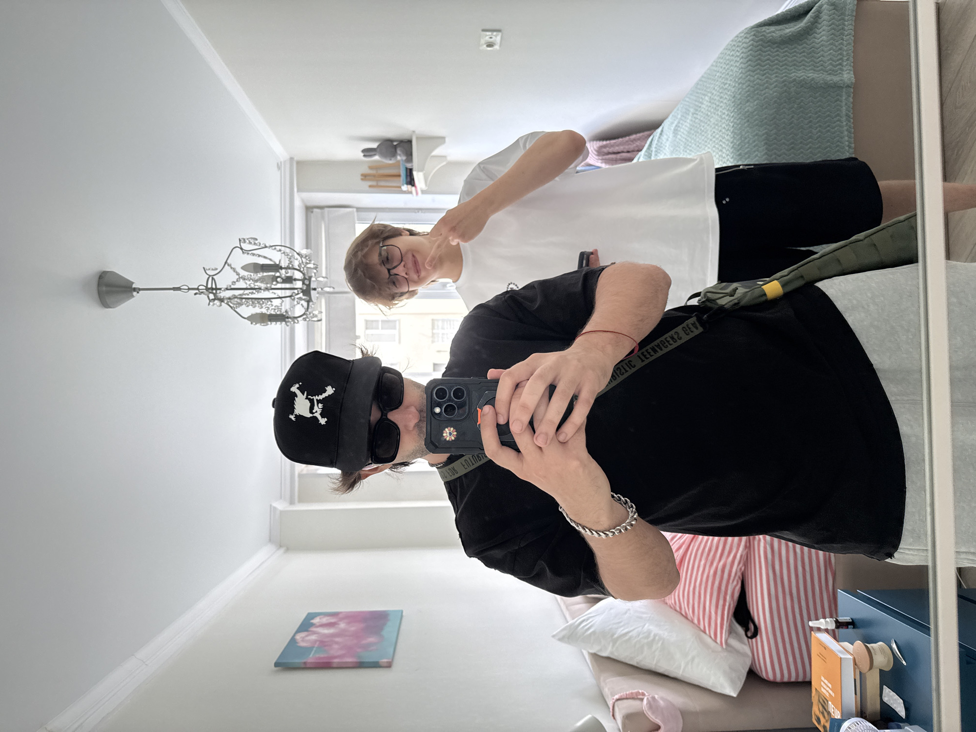 Two young men are reflected in a mirror in what appears to be a bedroom in Moscow, Russia. The man on the left is wearing a black t-shirt, grey sweatpants, a black baseball cap with a white skull emblem, and sunglasses. He is holding a smartphone, taking a selfie. He has a silver chain bracelet on his left wrist. A dark green messenger bag with a yellow strap is slung across his body. The man on the right is wearing a white t-shirt and black shorts. He is wearing glasses and has his right hand raised, fingers extended in a peace sign. In the background, a bed with striped pillows and a light green cover is visible. A bedside table with books and a small bottle is to the left. A chandelier hangs from the ceiling. The overall impression is that of a casual, personal moment captured in an indoor setting.