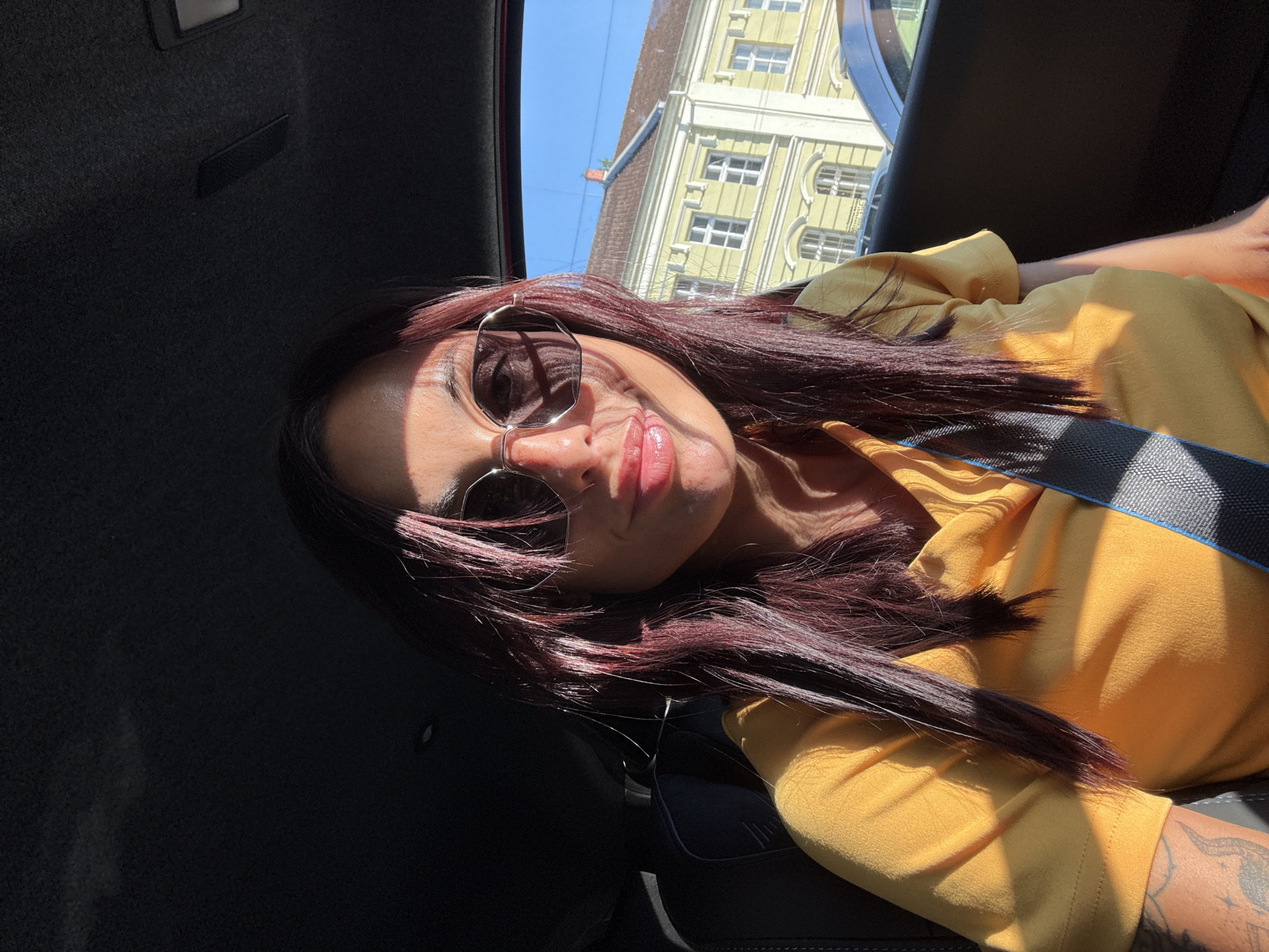 A woman is seated in the driver's side of a car, looking at the camera. She is wearing a yellow t-shirt and sunglasses. The interior of the car is dark, with sunlight coming in from the right side, casting shadows on her face and hair. Outside the car window, a yellow building with white window frames can be seen under a clear blue sky. The building is likely located in Pleven, Bulgaria, a city known for its historic architecture and sunny climate. The woman's arm, visible on the left side of the frame, has tattoos. The seatbelt is visible across her chest.