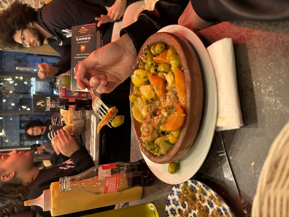 The image depicts an interior scene within a Numidia restaurant in Paris, France, likely during the evening. In the foreground, a brown clay tagine contains a meal featuring meat, potatoes, green olives, and orange vegetables, served on a white plate over a folded white napkin. A hand holds a silver fork, laden with a piece of meat and two green olives, poised above the tagine. Adjacent to the tagine are a small patterned plate holding a green relish and a 250ml glass bottle of Coca-Cola, alongside a large yellow squeeze bottle.
In the mid-ground, three individuals are seated at a dark table. On the left, a young female with dreadlocks looks to the right. To her right, a young male with an afro and glasses holds a white smartphone vertically. In the background, another person with curly hair and glasses is visible, smiling. The table also holds another glass of dark liquid, a red can, and a menu board displaying "NUMIDIA" and a Paris phone number (01 72 00 38 79). The restaurant's name and logo are also visible on an illuminated sign through a window in the background. The environment features dark tables, a brick wall to the right, and soft, decorative lighting in the background.