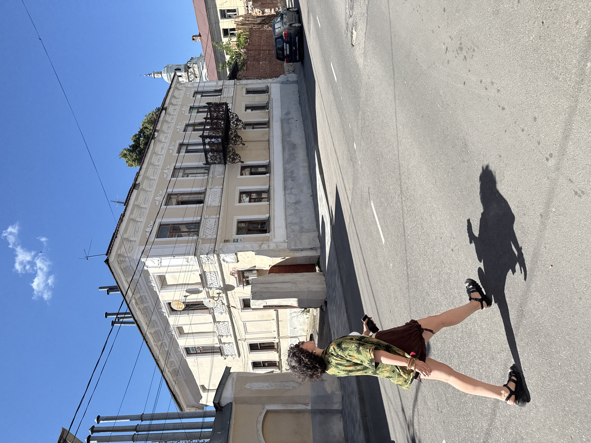 In Yelets, Russia, under a bright blue sky with a few wispy clouds, a person with curly dark hair walks across a street. They are wearing sunglasses, a green shirt with a tropical print, and a brown skirt. Black sandals are on their feet, and they are holding a dark-colored smartphone in their right hand. The person casts a long shadow on the asphalt road, which has white lane markings.
Behind the person is a street sidewalk and a light-colored, multi-story building with ornate architectural details. The building has several windows, some with brown frames. A black wrought-iron balcony juts out from the upper floor. Adjacent to this building is another structure, partially visible, with tall, grey, cylindrical chimney-like objects protruding from its roof. Further in the background, other buildings are visible, including one with a prominent white dome and spire, suggesting it might be a church or cathedral. A dark-colored SUV is parked on the right side of the street, near a brick wall. The overall scene depicts a sunny day in an urban environment.