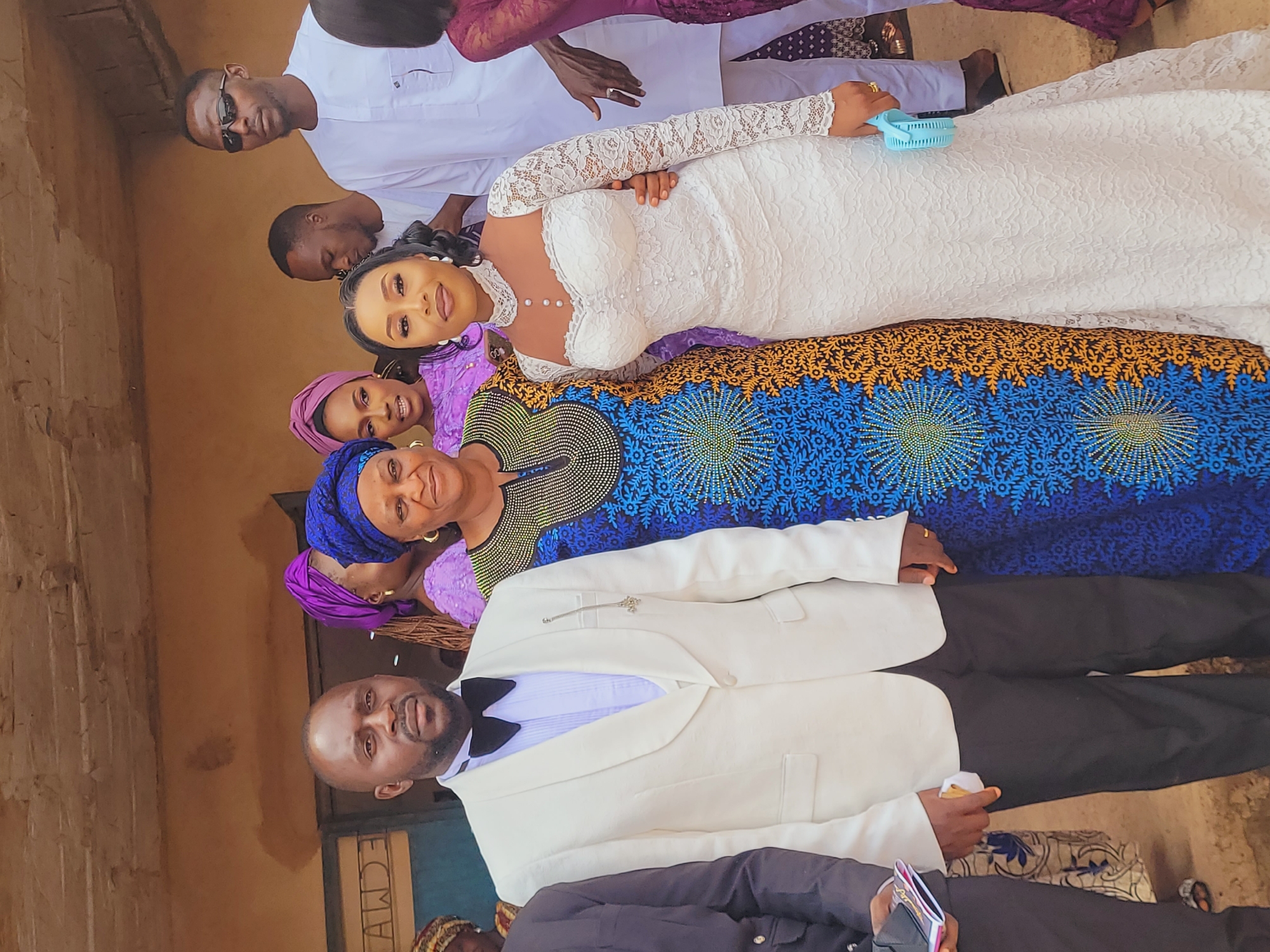 A group of individuals is gathered outdoors in Bukuru, Nigeria, in front of a tan-colored building with visible signs of wear and discoloration. The foreground features three central figures: a man, and two women. The man on the left, an African male with a short beard, wears a cream-colored tuxedo jacket over a white pleated shirt with a black bow tie and dark trousers. A silver chain ornament is pinned to his left lapel. To his right stands an African woman in a vibrant blue headwrap and a patterned blue, orange, and black gown, adorned with small beads on the neckline. Her left arm is placed around the waist of the woman to her right. This woman, an African female with a lighter complexion, wears a form-fitting white lace wedding gown with long sleeves, a high lace choker neckline accented with pearls, and pearls running down the front of the bodice. She holds a light blue portable fan in her right hand and has her left arm around the back of the woman in the blue dress. All three central figures look towards the camera.
In the background, several other people are present. On the far right, an African male in a white traditional Nigerian outfit and dark sunglasses stands looking left. Another man in a white shirt is partially visible behind the woman in the white dress. Two women, one partially visible, are positioned behind the central women, both wearing purple headwraps and patterned purple dresses. On the far left, a person in a dark suit is partially visible. The background building wall is uneven, and a blue door or window frame on the left shows partial, possibly reversed, lettering. The ground beneath the individuals is unpaved and light brown, with a raised edge or step visible.