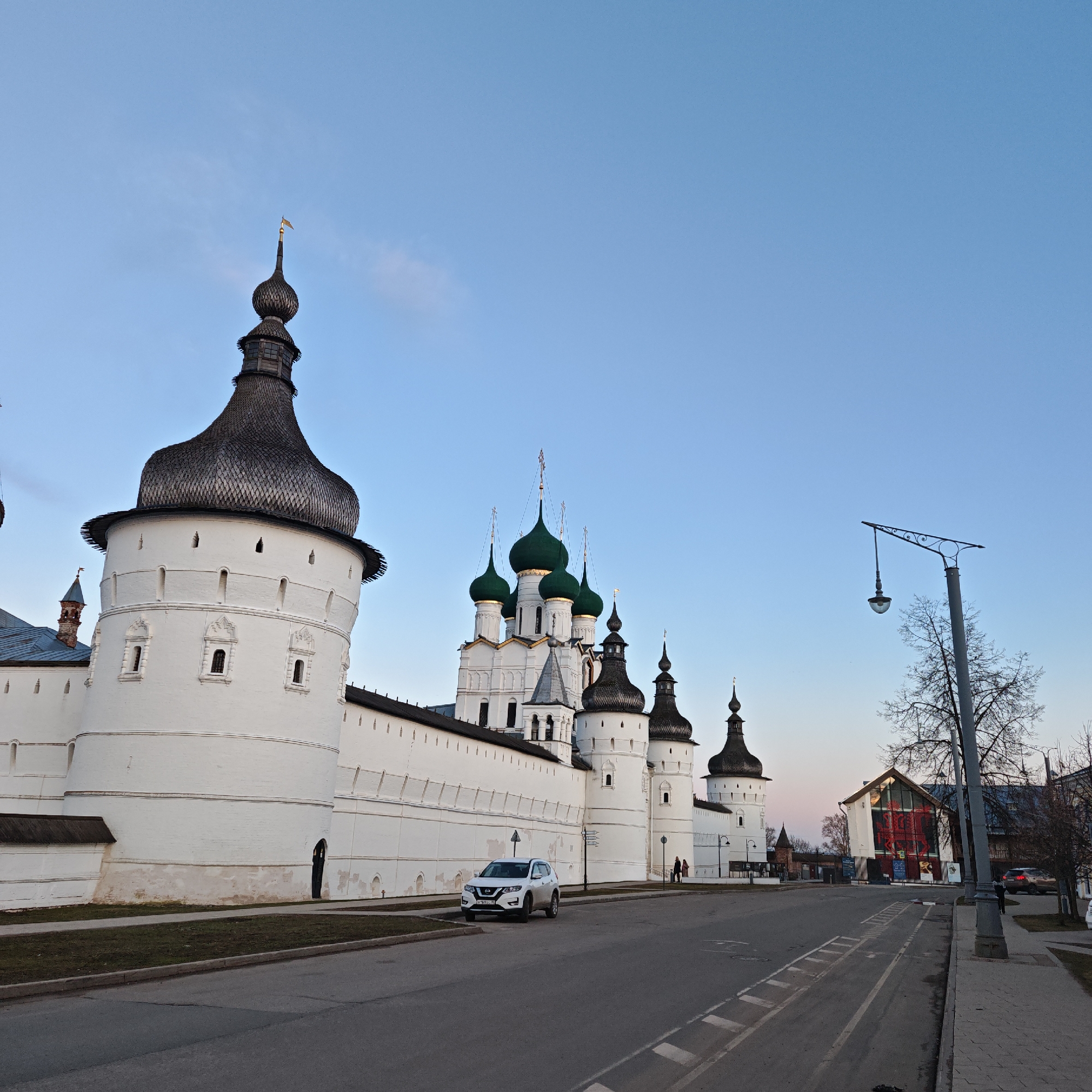 The image depicts a street scene in Rostov, Russia, featuring a section of the Rostov Kremlin. The Kremlin wall and towers are prominent, painted white with dark, distinctive roof structures. A modern white SUV is parked near the wall. The road is asphalt, with painted white markings. A lamppost and bare tree are on the right side of the image, near a sidewalk. A building with a large graphic design and multiple windows is visible in the background. The sky is clear with a gradient of light blue and pink, indicating either sunrise or sunset.