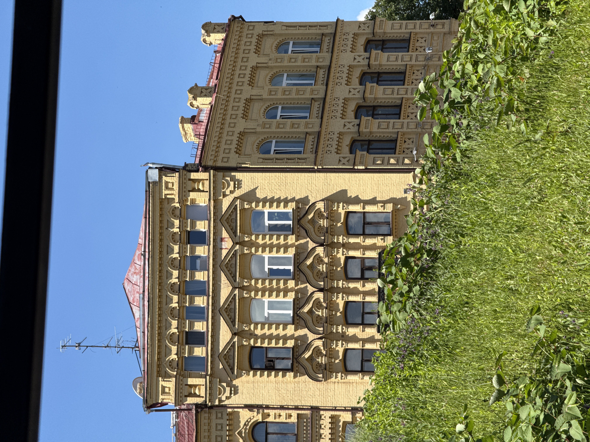 This is a close-up, eye-level, outdoor, daytime, medium shot of a yellow brick building in Kyiv, Ukraine. The building has a red roof and many decorative architectural elements, including patterned brickwork, arched windows, and ornate window frames. In the foreground, a thick patch of green grass and other vegetation, some with small purple flowers, obscures the lower portion of the building. A black horizontal bar runs across the top of the image, suggesting the photo was taken through a window or from behind an obstruction. The sky is a clear, bright blue. There are no people visible in the image.