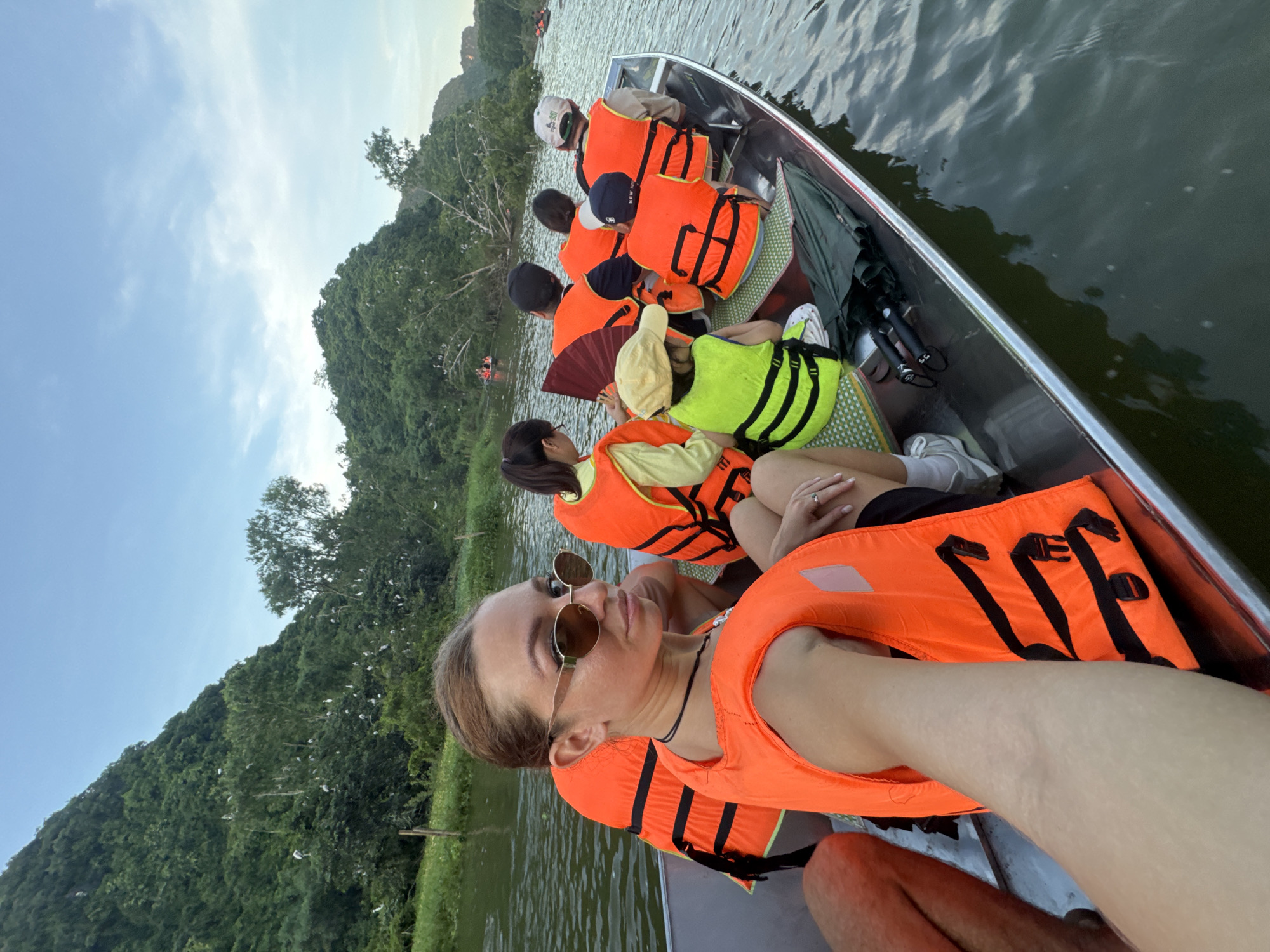 In Yên Sơn Ward, Vietnam, a group of people are on a boat tour on a river, surrounded by lush green hills and trees. The water is calm and reflects the sky. Several individuals are wearing orange life vests, suggesting a tourist activity. In the foreground, a woman is taking a selfie, looking at the camera with sunglasses on. She is wearing an orange life vest. Other people in the boat are facing away from the camera, looking at the scenery. Some are children wearing bright green vests. There are birds visible in the trees on the hillsides. The overall scene depicts a leisurely boat excursion in a natural setting.