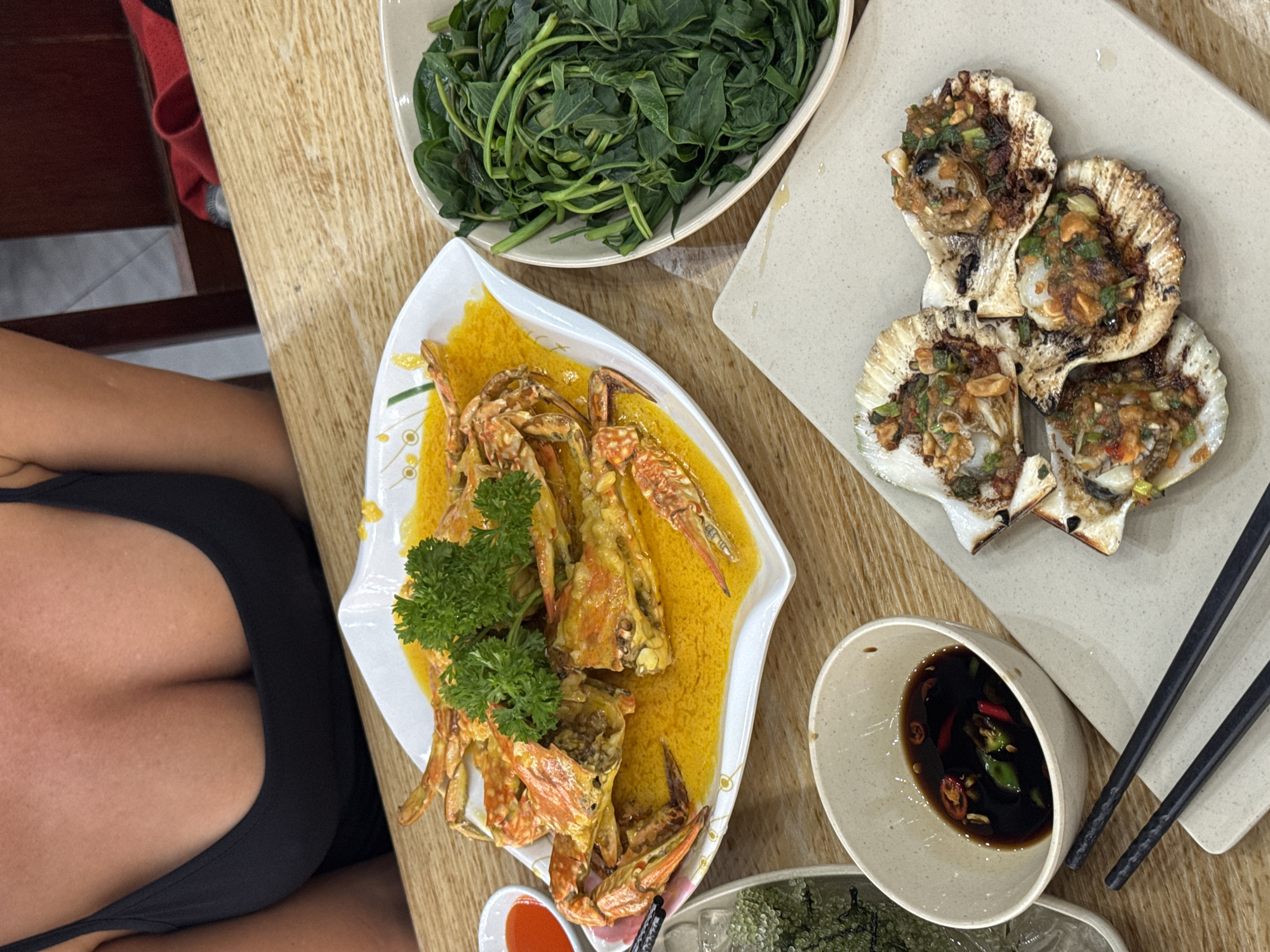 A top-down, eye-level shot reveals a dining table laden with Vietnamese cuisine. To the left of the frame, a person is partially visible, wearing a black top. The table is adorned with several dishes: a white bowl filled with leafy green vegetables, a plate of crab in a yellow sauce garnished with parsley, a square plate with scallops topped with herbs and spices, and a small bowl containing a dark sauce with sliced chili peppers. Black chopsticks rest on the square plate. The scene appears to be set in Đà Nẵng, Vietnam, evidenced by the style of dishes commonly found in the region.