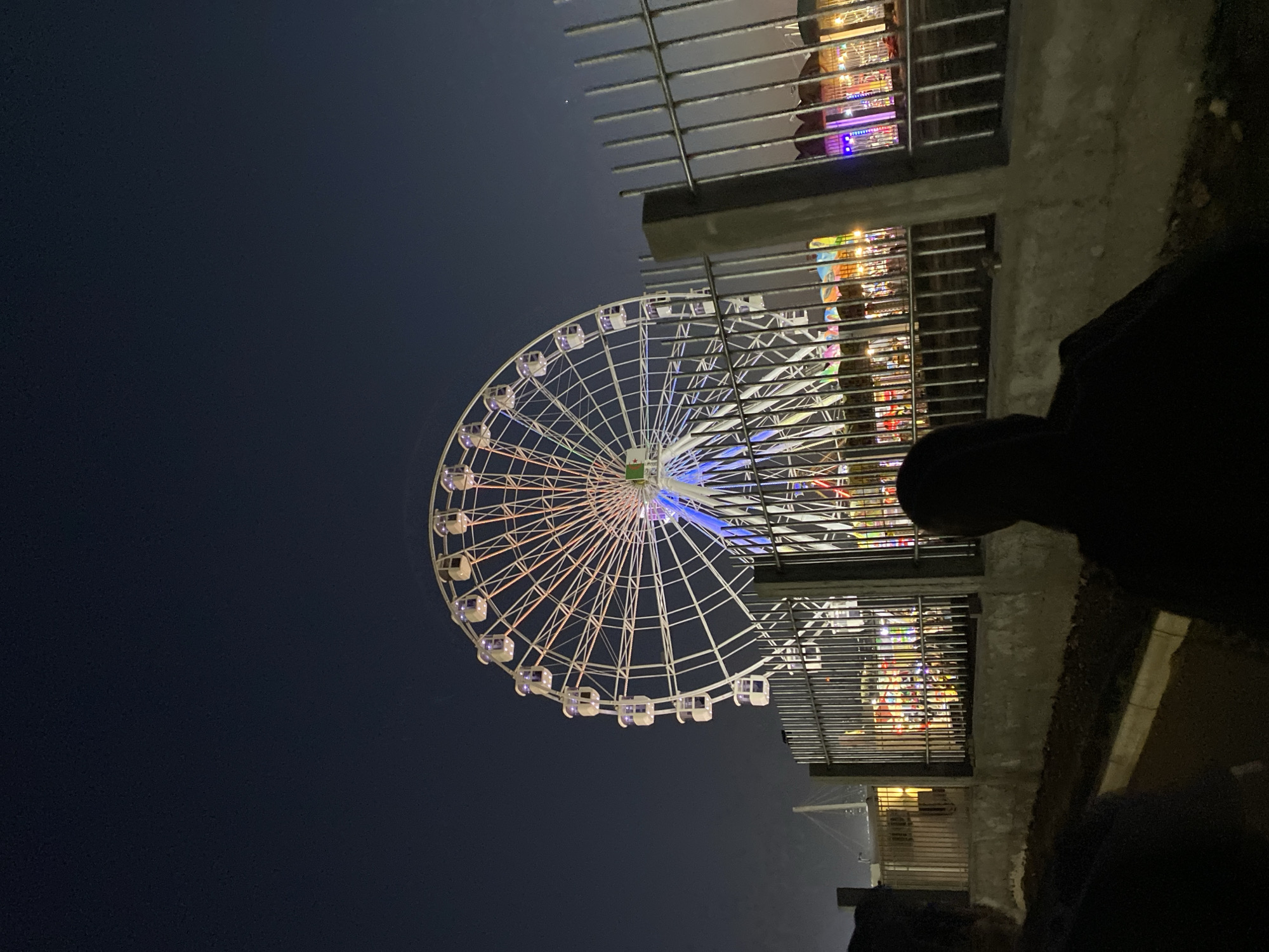 The image shows a Ferris wheel at night, likely in Hussein Dey, Algeria. The Ferris wheel is brightly lit with white, blue, and red lights, and it appears to be the main attraction of a fair or amusement park. In the foreground, there is a metal fence, and behind it, there are some structures that seem to be part of the amusement park, possibly rides or stalls, also illuminated by colorful lights. The sky is dark, indicating nighttime. Two individuals are silhouetted in the foreground, facing away from the camera and towards the Ferris wheel and the park. The ground in the foreground appears to be a pathway or road.