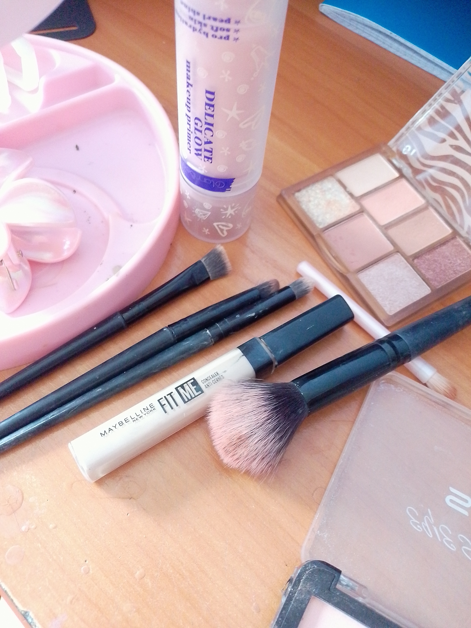 A close-up, overhead shot displays a collection of makeup items arranged on a wooden surface. A pink plastic makeup organizer is visible on the left, holding several brushes. In the center, a white tube of Maybelline Fit Me concealer lies next to a makeup palette with various eyeshadow shades. A fluffy pink and black makeup brush rests diagonally across the image, its bristles fanned out. To the right, a translucent compact with a zebra-patterned lid contains more makeup. The lighting is bright, casting soft shadows. The scene appears to be an indoor setting, possibly a dressing table or vanity. There is no identifiable location in the city of Pryluky, Ukraine.