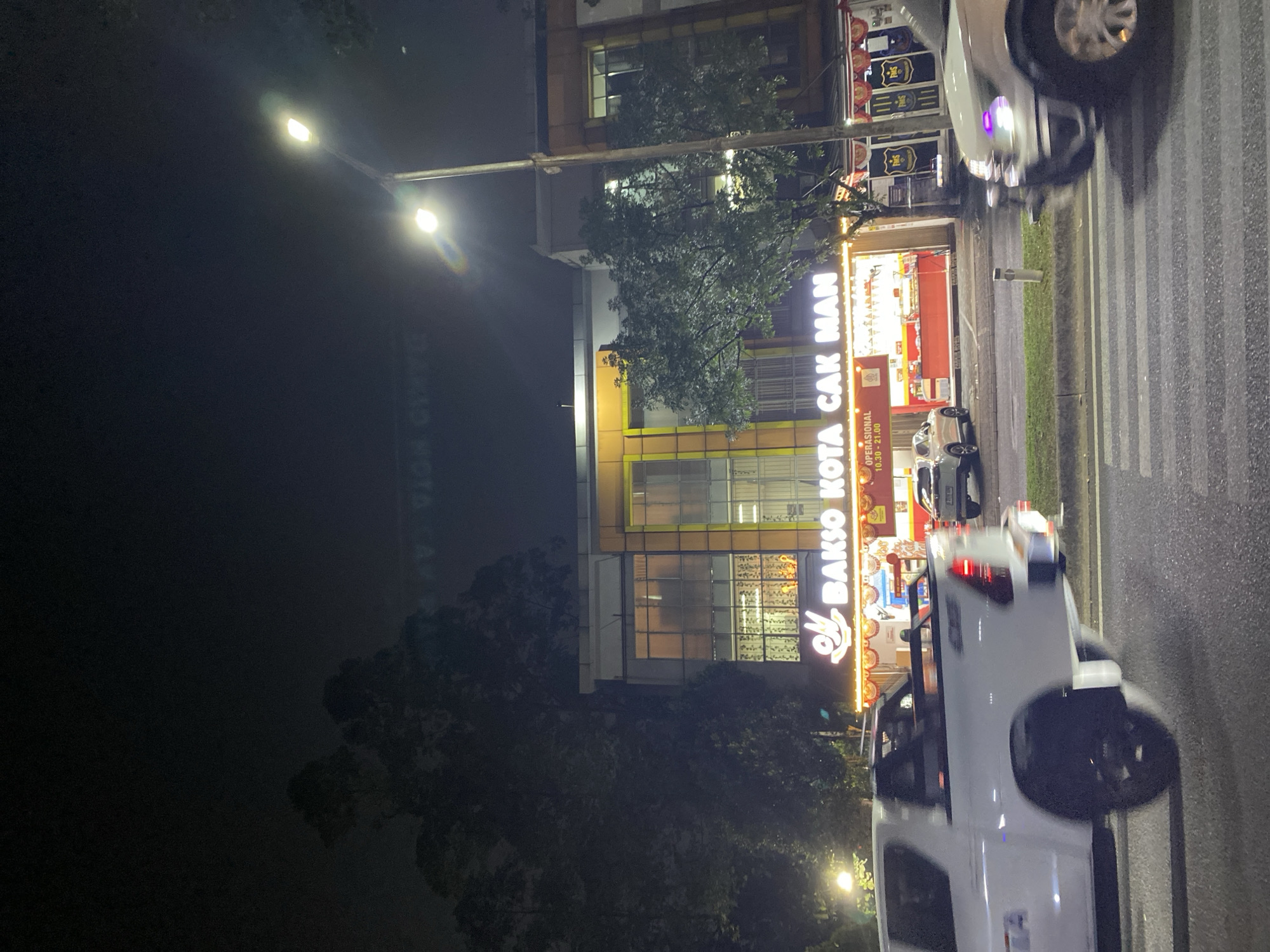 This is a nighttime outdoor, medium-distance photograph of a street scene in South Tangerang, Indonesia. In the foreground, a white pickup truck is captured in motion, with its rear right tire and fender prominently displayed. To the right of the pickup truck, the front of another white vehicle is visible, with its headlight illuminated and a purple glare on its front bumper. The middle ground features a building with bright signage that reads "BAKSO KOTA CAK MAN." Below the sign, a food stall with red and yellow accents is visible, displaying various food items. The stall's operating hours, "OPERASIONAL 10.30 - 21.00," are also displayed. Trees are present on the left and right sides of the building, and a streetlamp with two illuminated bulbs is visible on the right side of the frame. The background is dark and obscured by trees and foliage.