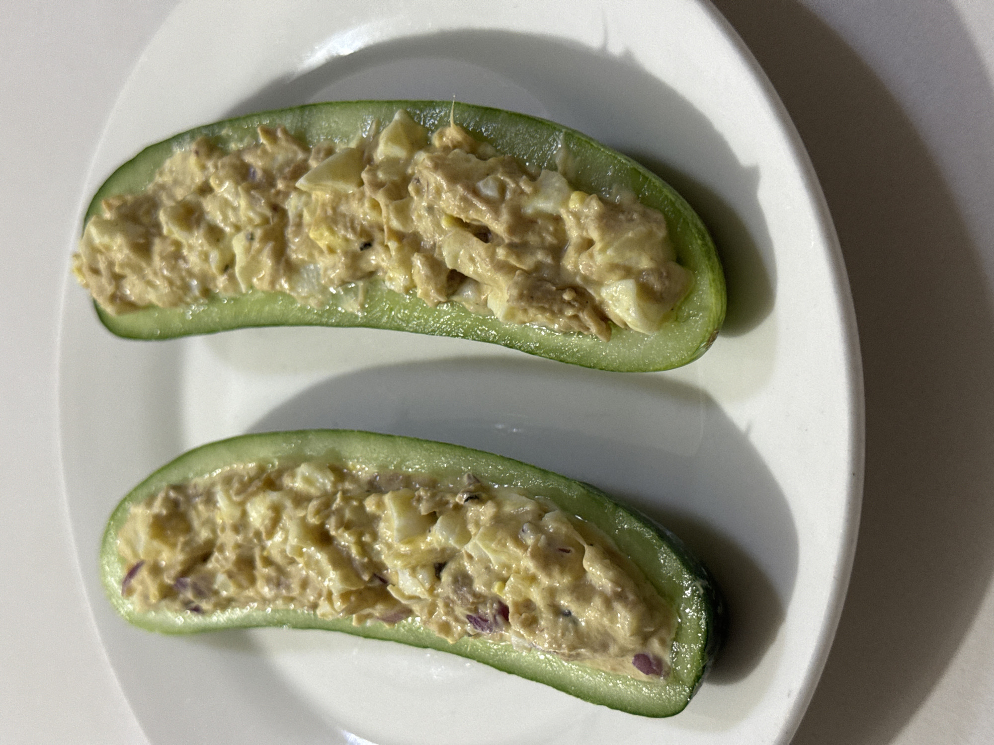 Two halves of a cucumber, hollowed out and filled with a mixture of tuna, chopped hard-boiled eggs, and possibly red onion, rest on a white plate. The plate is positioned on a neutral-colored surface, casting soft shadows. The scene is captured from a top-down perspective. This image does not contain any information related to Davao City, Philippines.